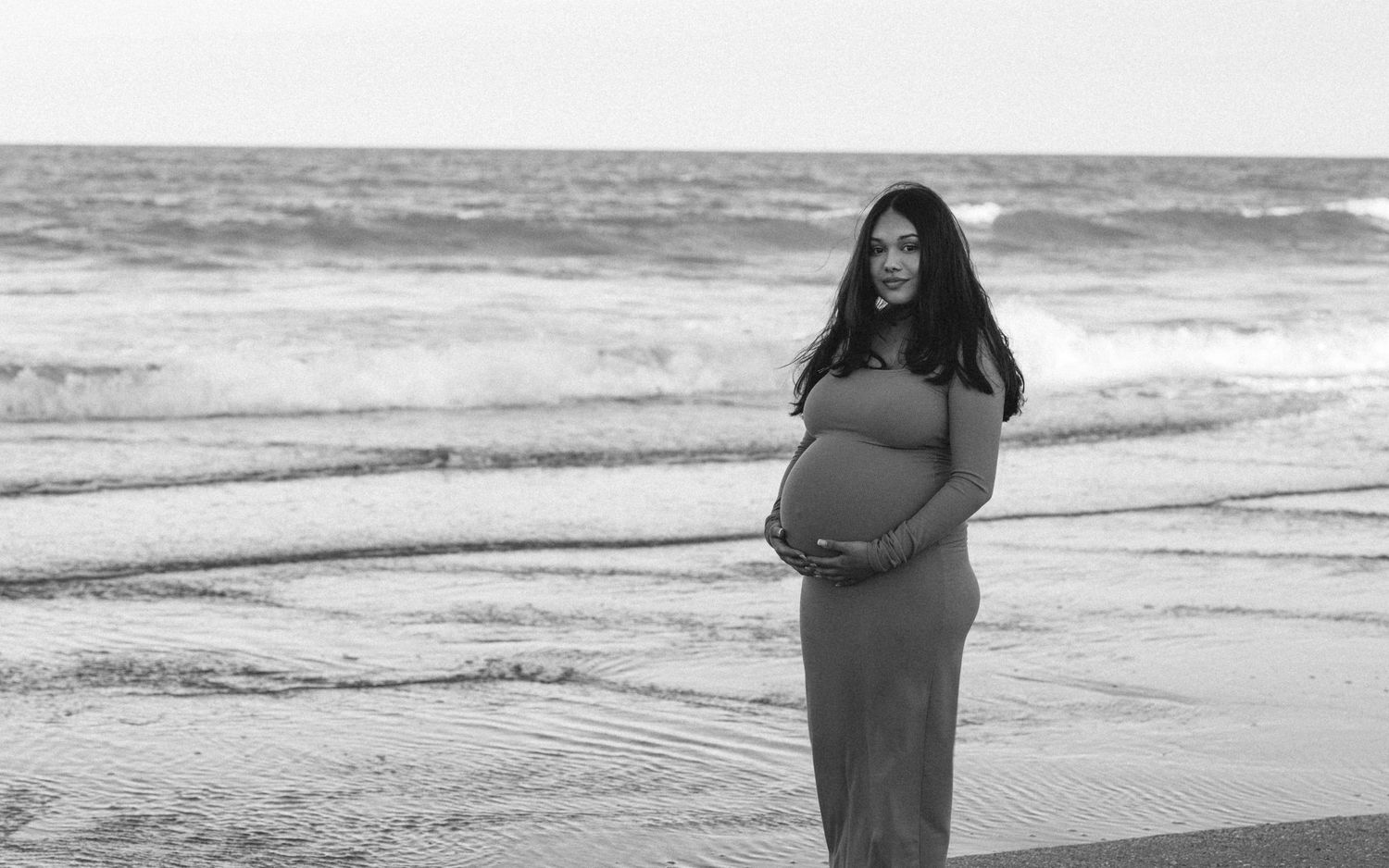 Black and white maternity photo of a pregnant woman standing on a beach holding her baby bump.