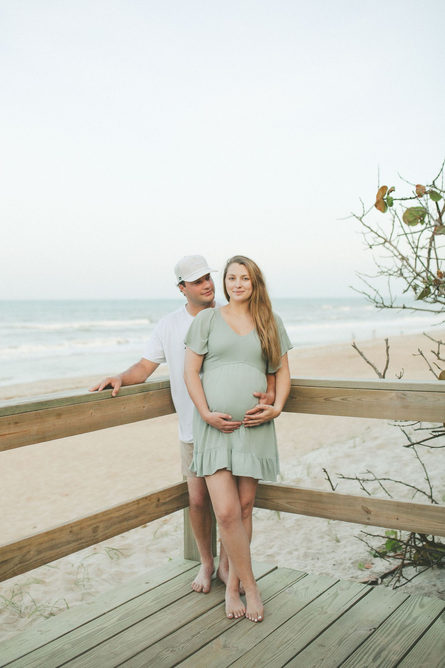 Pregnant couple posing on a beach boardwalk, woman in sage green dress cradling baby bump, ocean in background.