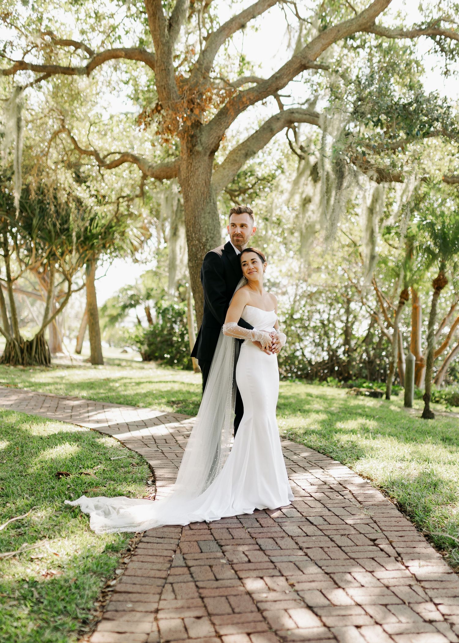 Bride and groom posing together on brick pathway under Spanish moss-draped oak trees.