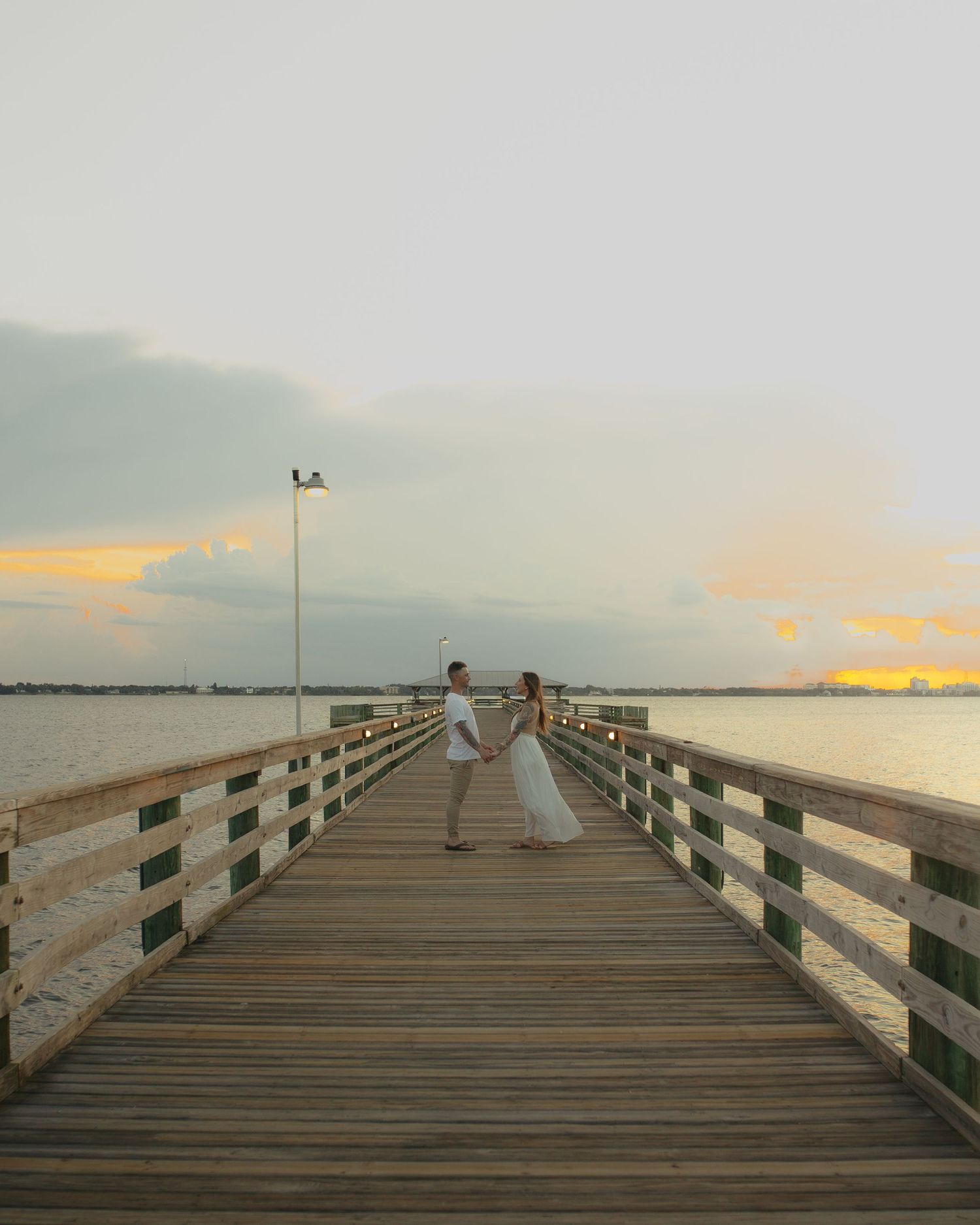 Romantic couple holding hands on a long wooden pier overlooking a calm bay at golden hour sunset.