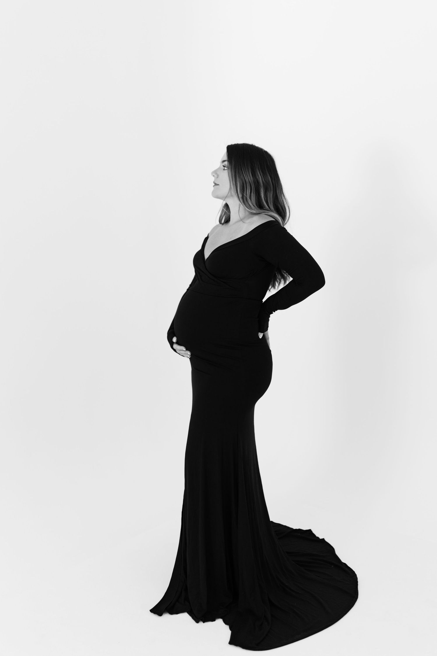 Elegant pregnant woman in a black off-shoulder gown posing sideways in a black and white studio photo.