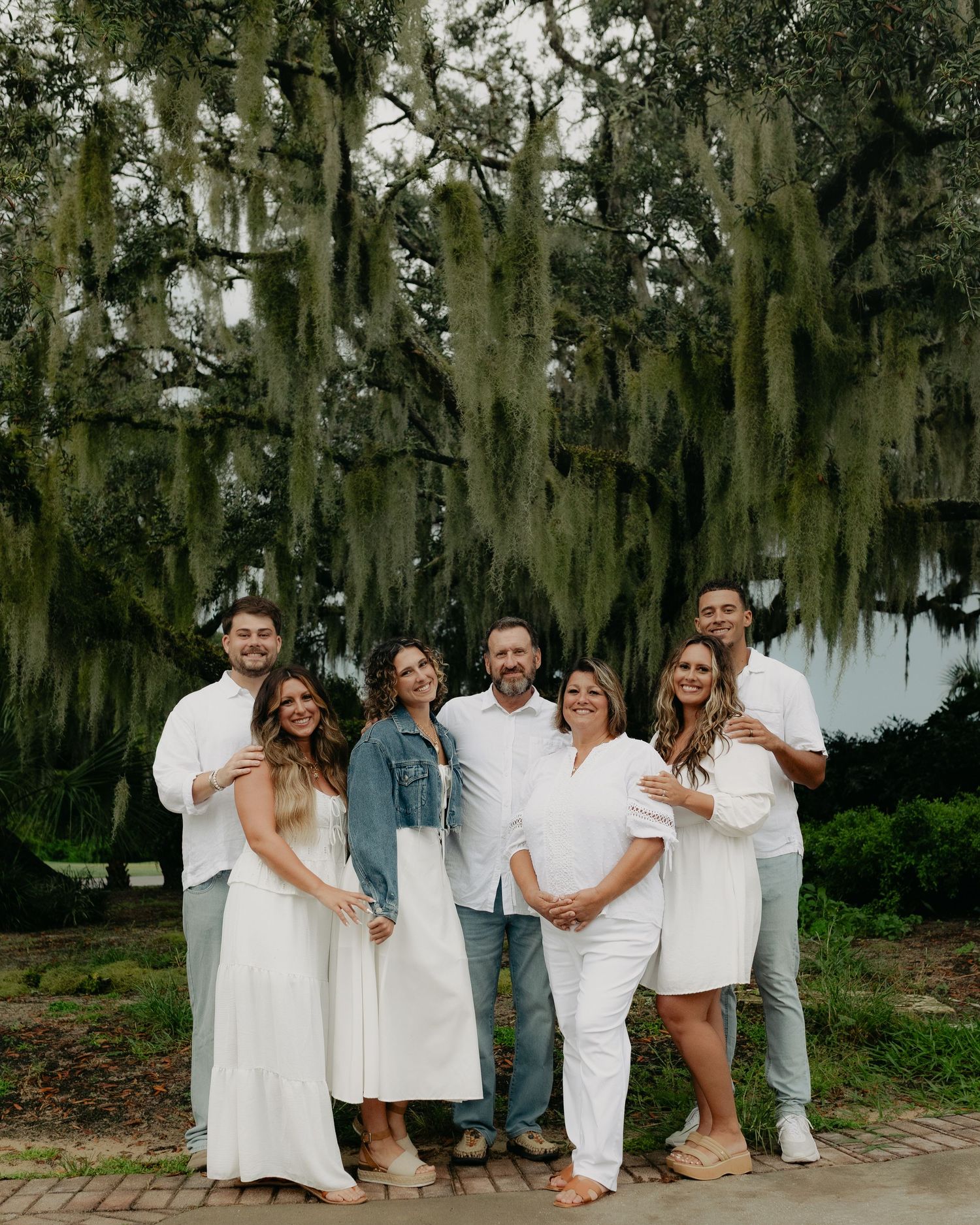 Seven-person family group smiling in white outfits under Spanish moss-covered oak trees.