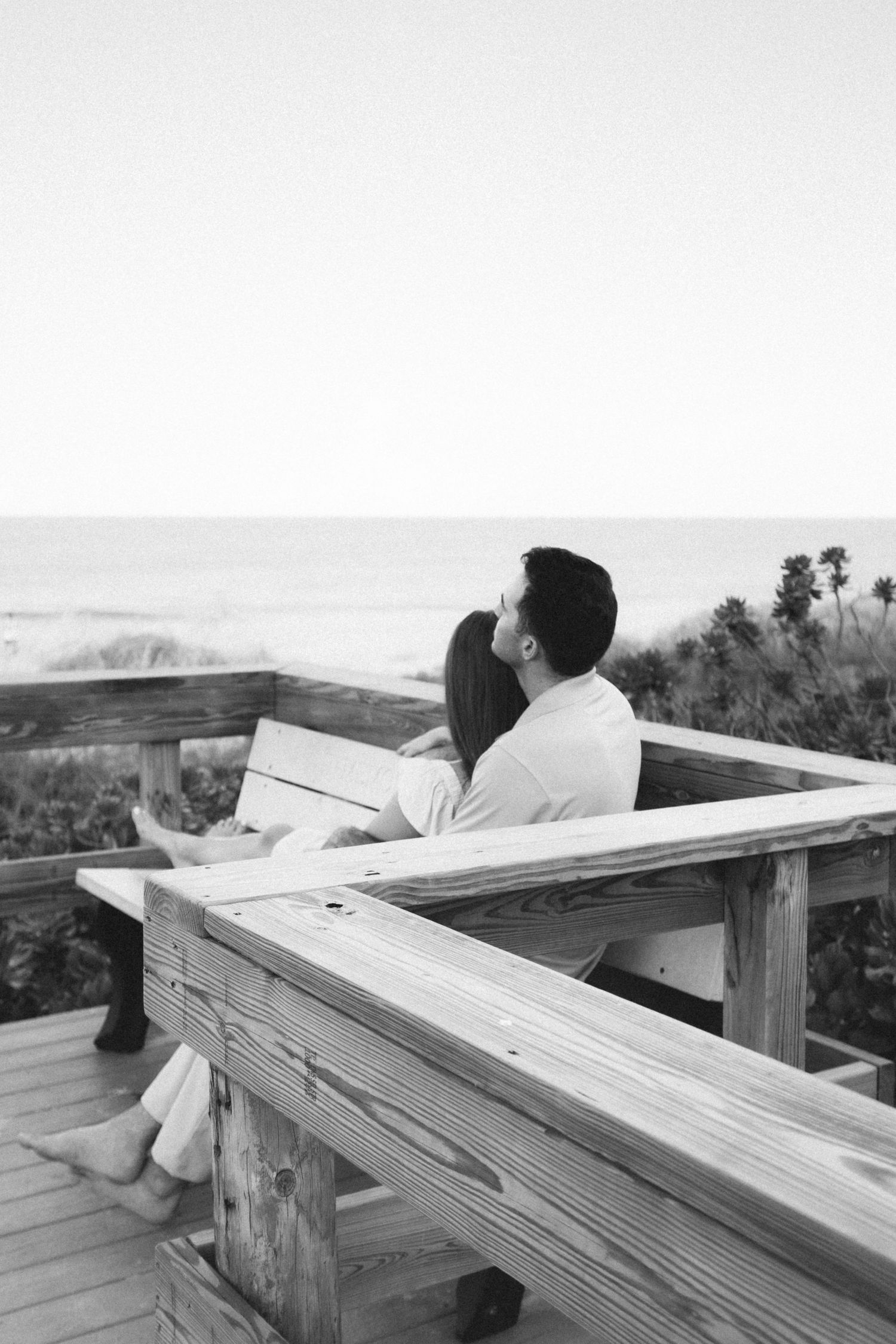 Black and white photo of a couple sitting together on a wooden deck bench overlooking the ocean.