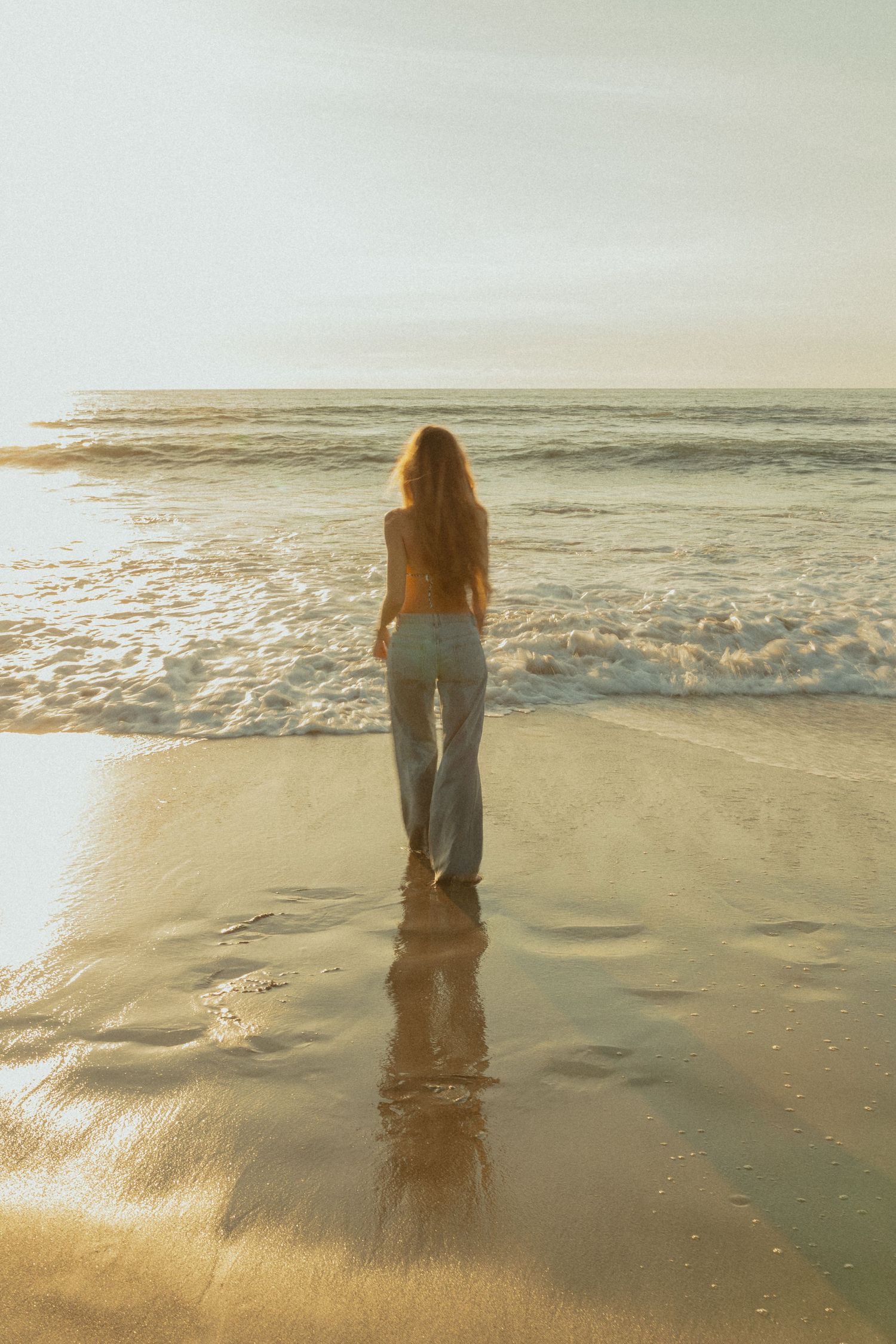 A woman with long hair stands barefoot on a wet sandy beach at golden hour, wearing light blue wide-leg pants.