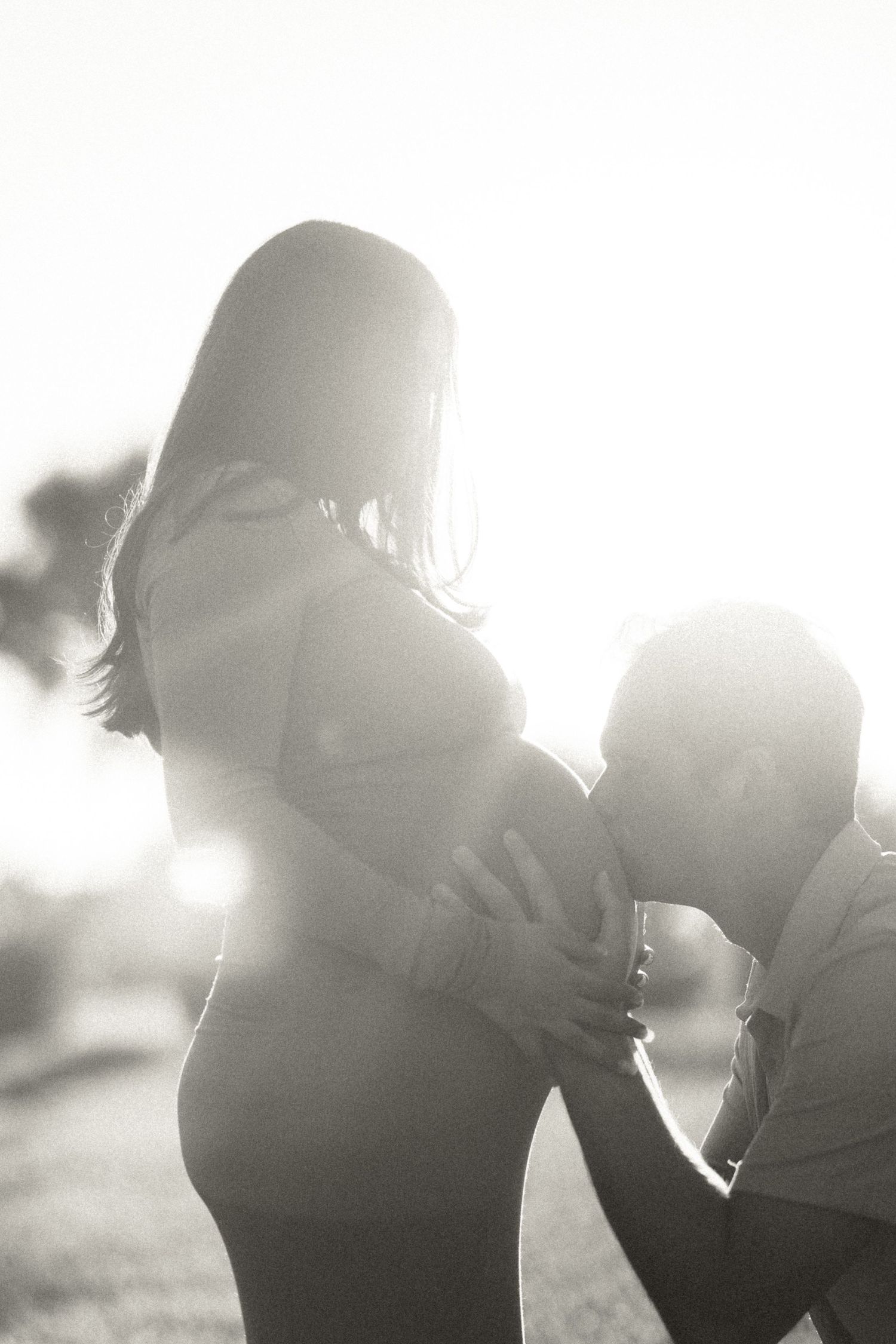 Backlit black and white silhouette of a man kissing a pregnant womans belly outdoors at sunset.