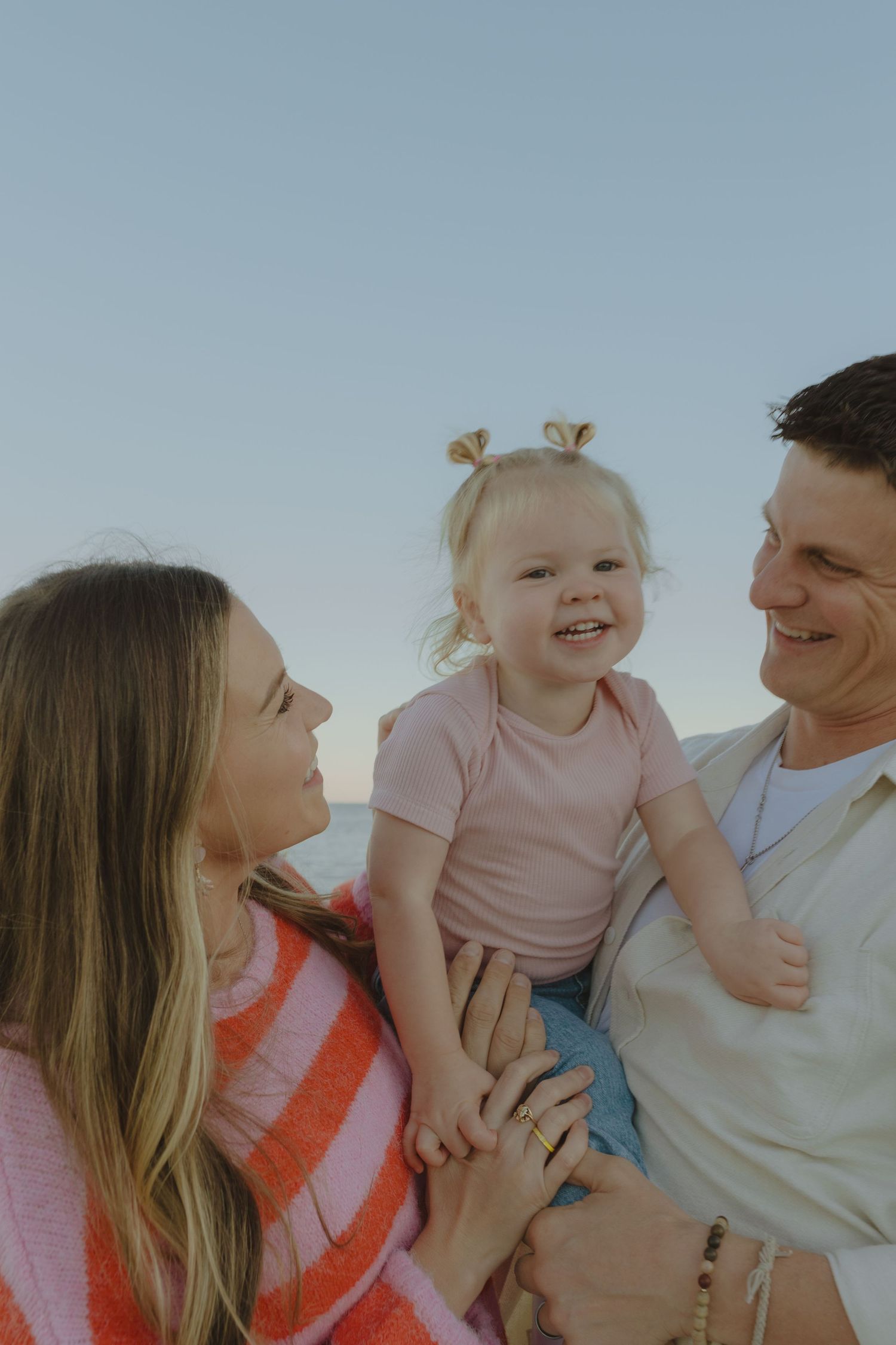 Joyful toddler with pigtails laughing between her smiling parents against a clear sky backdrop.