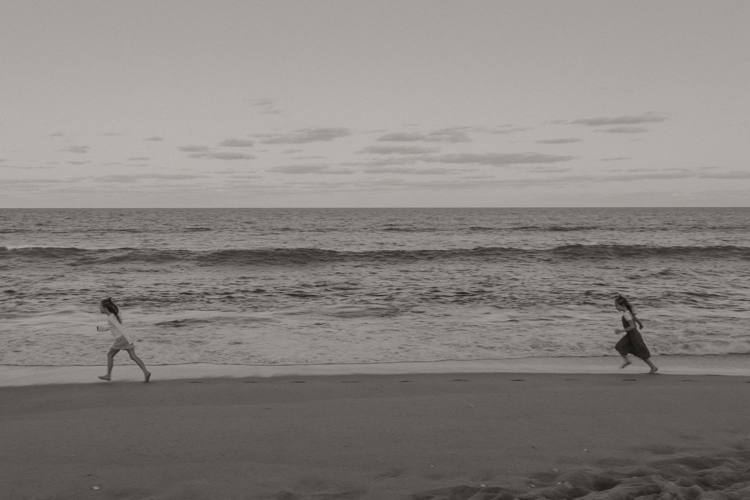 Black and white photo of two children running and playing along ocean shoreline at the beach.