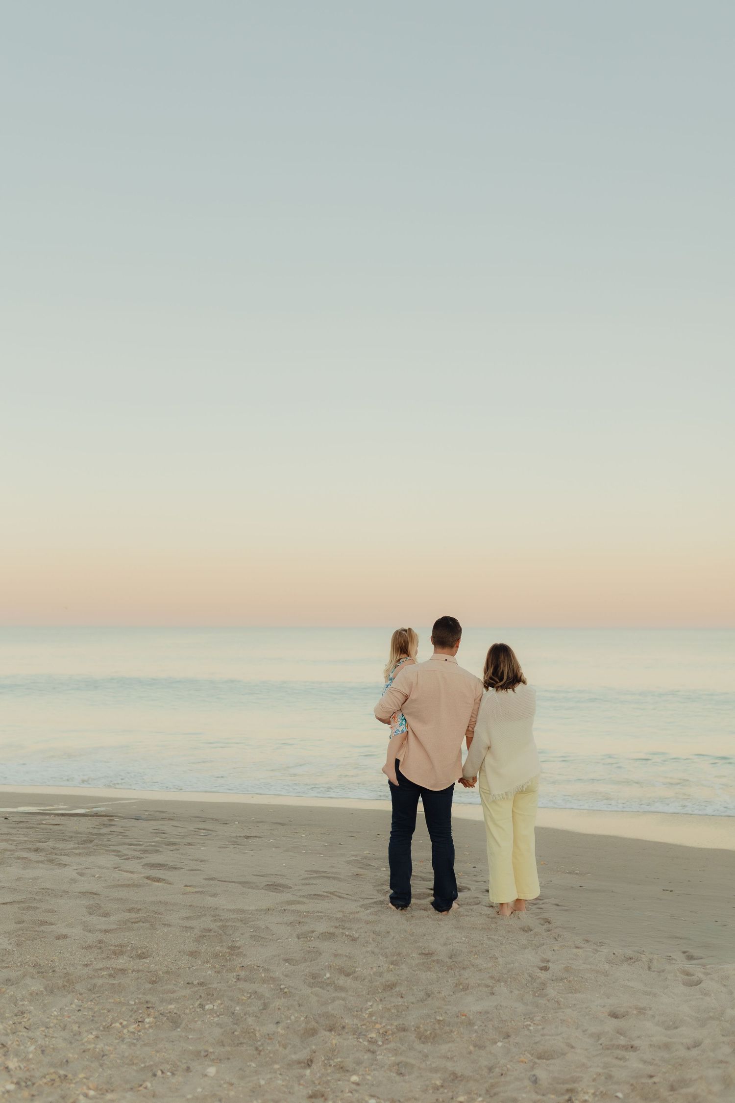 A family of three stands on a sandy beach at dusk, gazing at the calm ocean under a pastel sunset sky.