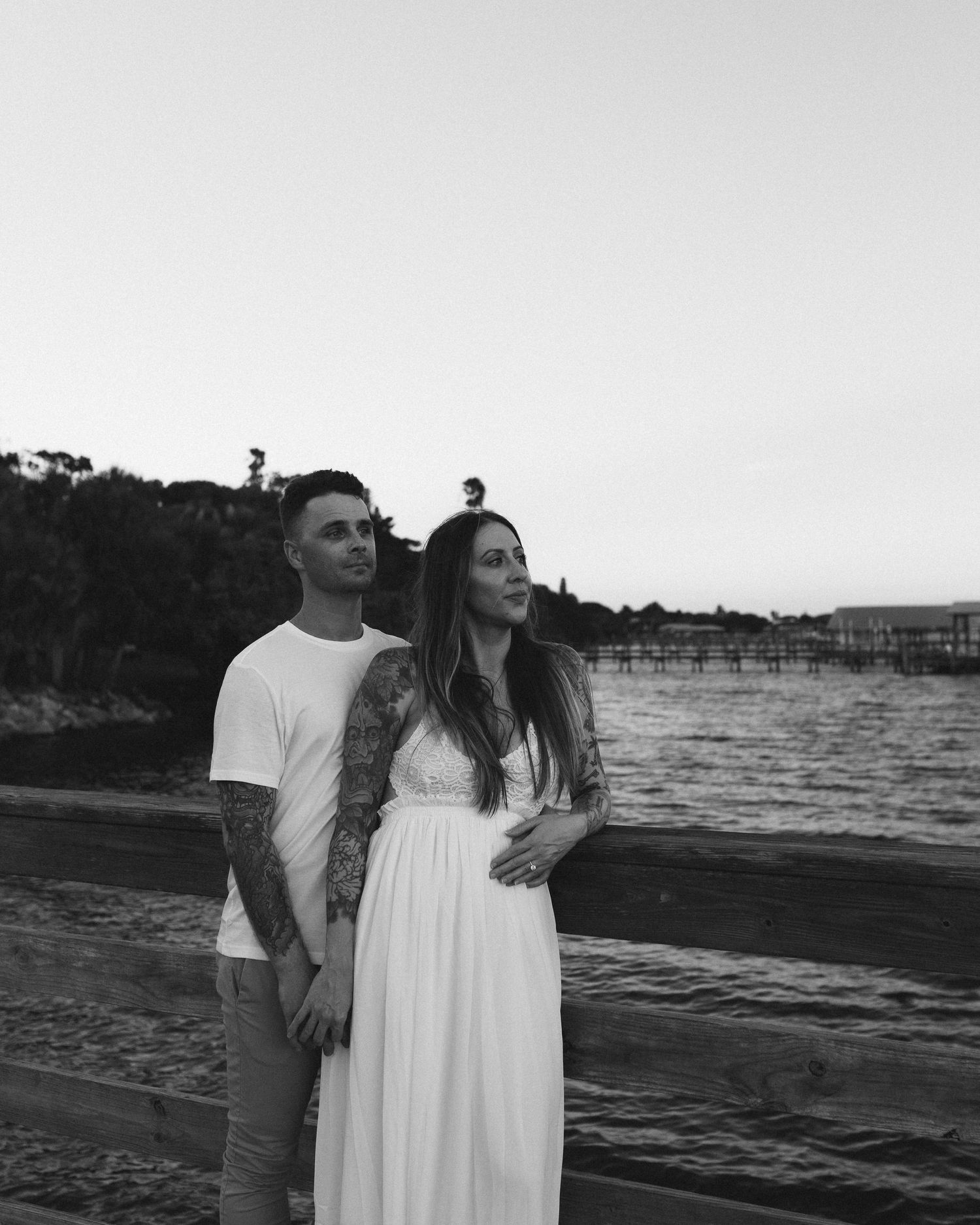 Black and white portrait of a tattooed couple standing together by the waterfront at sunset.