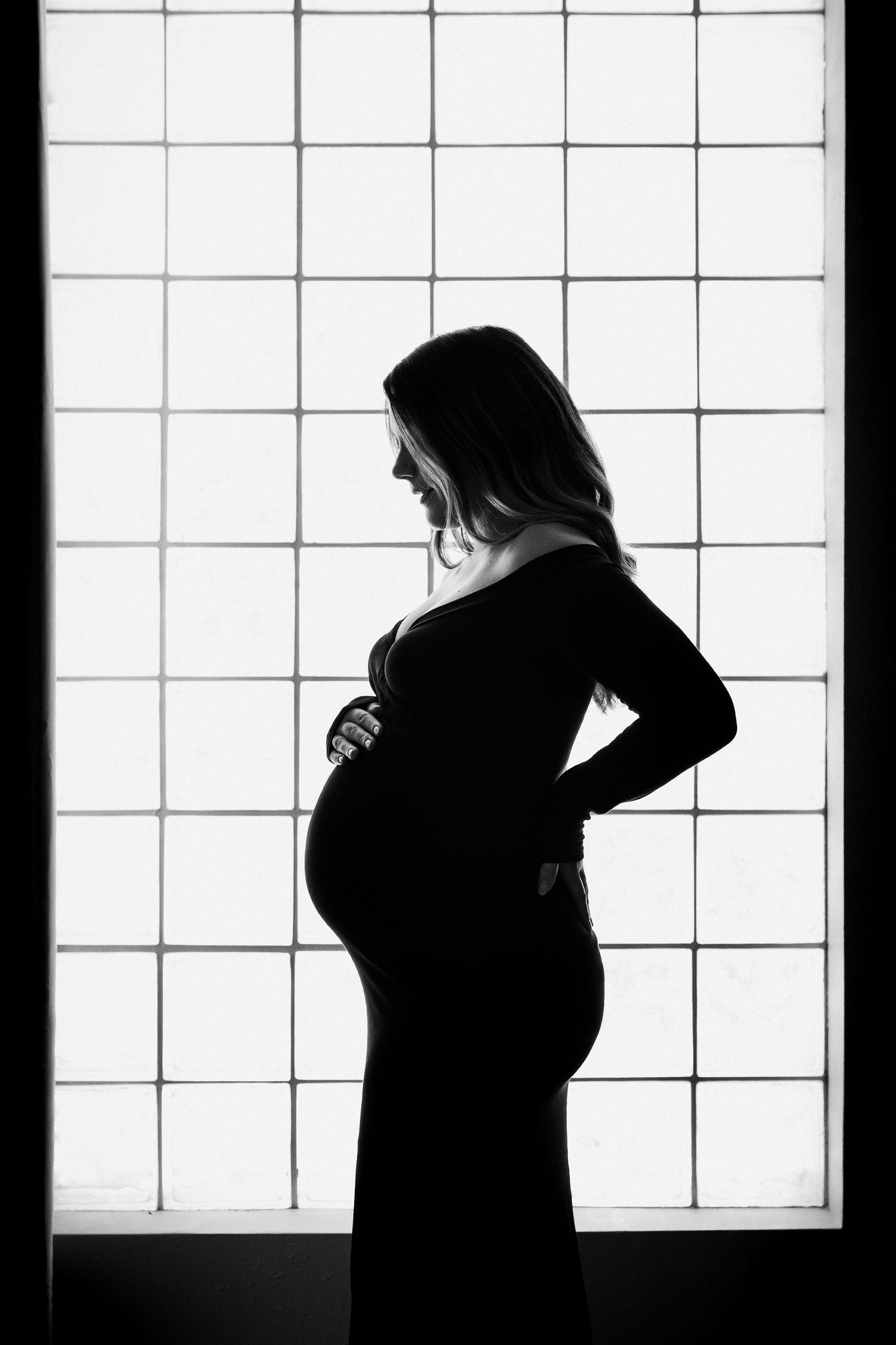 Striking black and white silhouette of a pregnant woman in a black dress posed before a grid window.