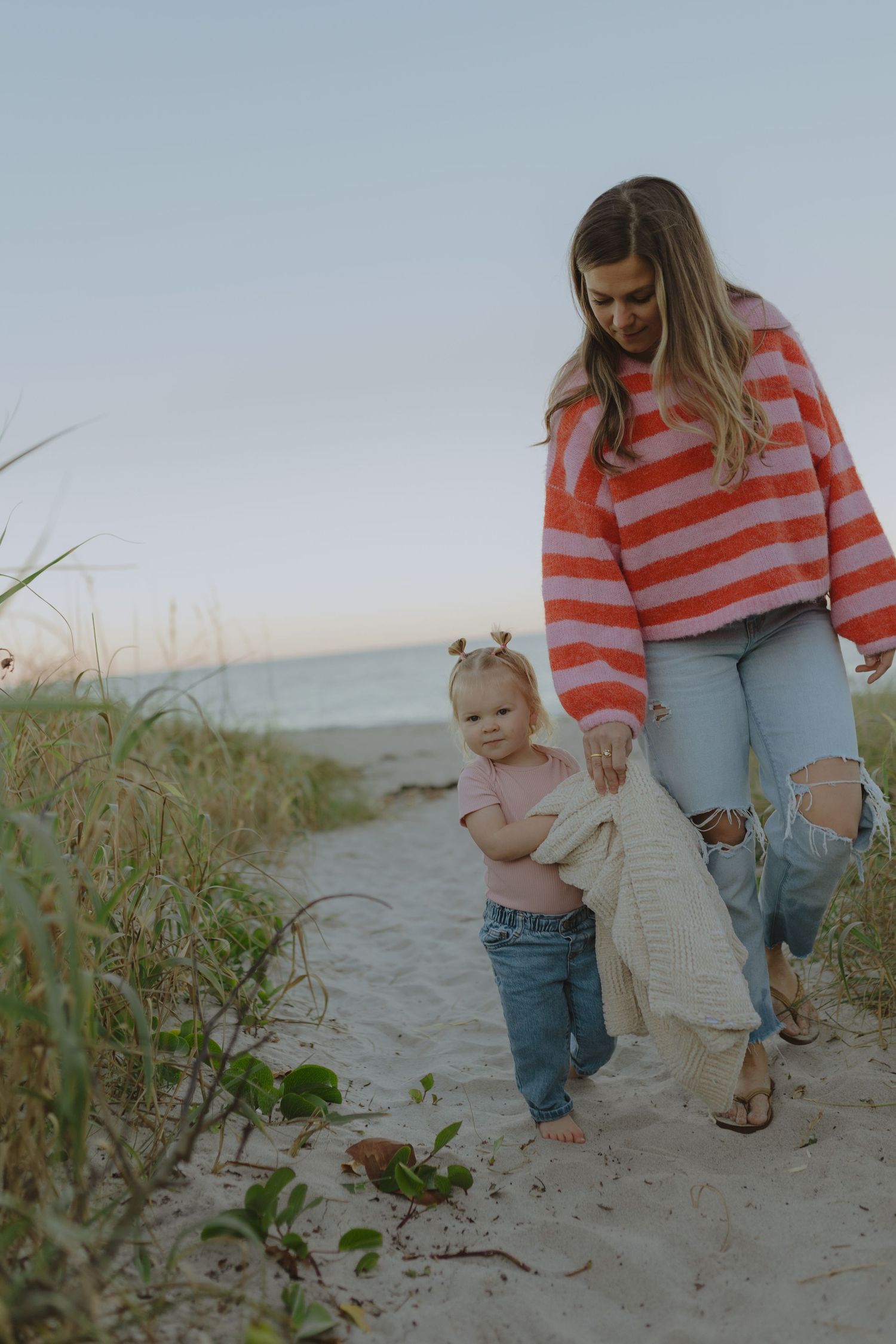 A mother in a striped sweater walks with her toddler on a sandy beach path at sunset.