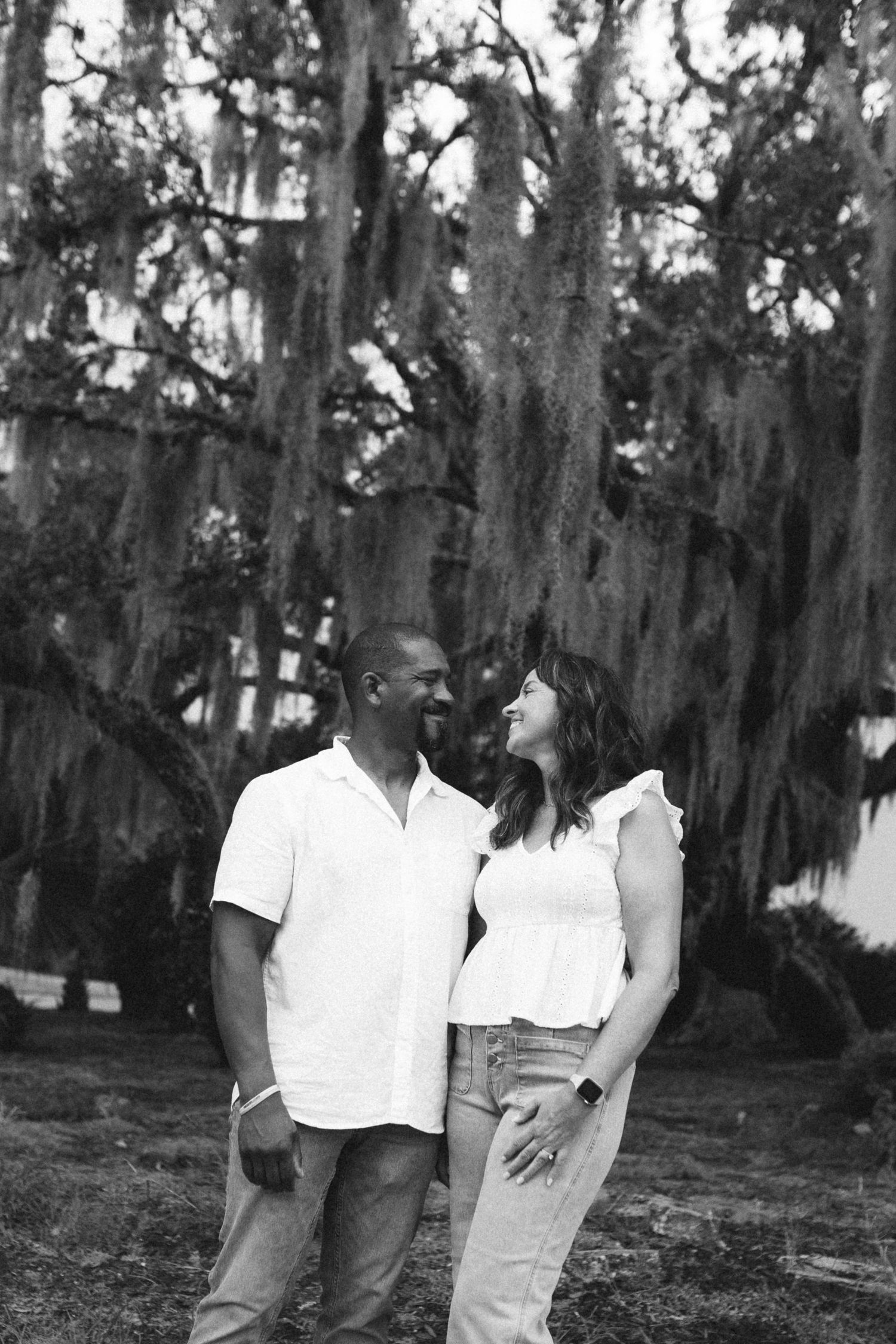 Black and white portrait of smiling couple in white outfits under Spanish moss trees.
