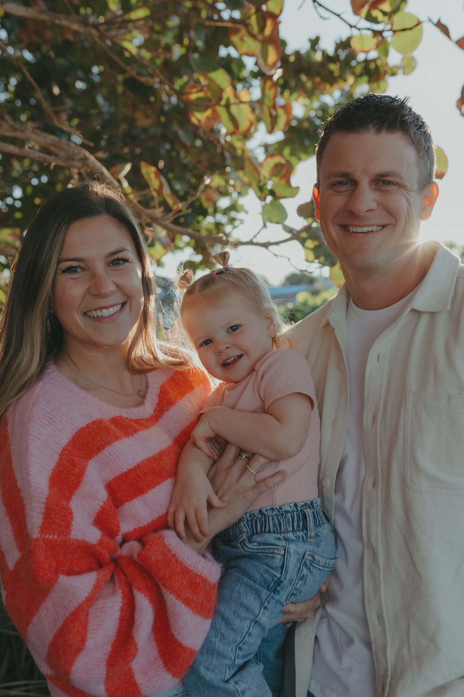 Smiling couple holding their laughing baby outdoors with sunlight filtering through green trees.