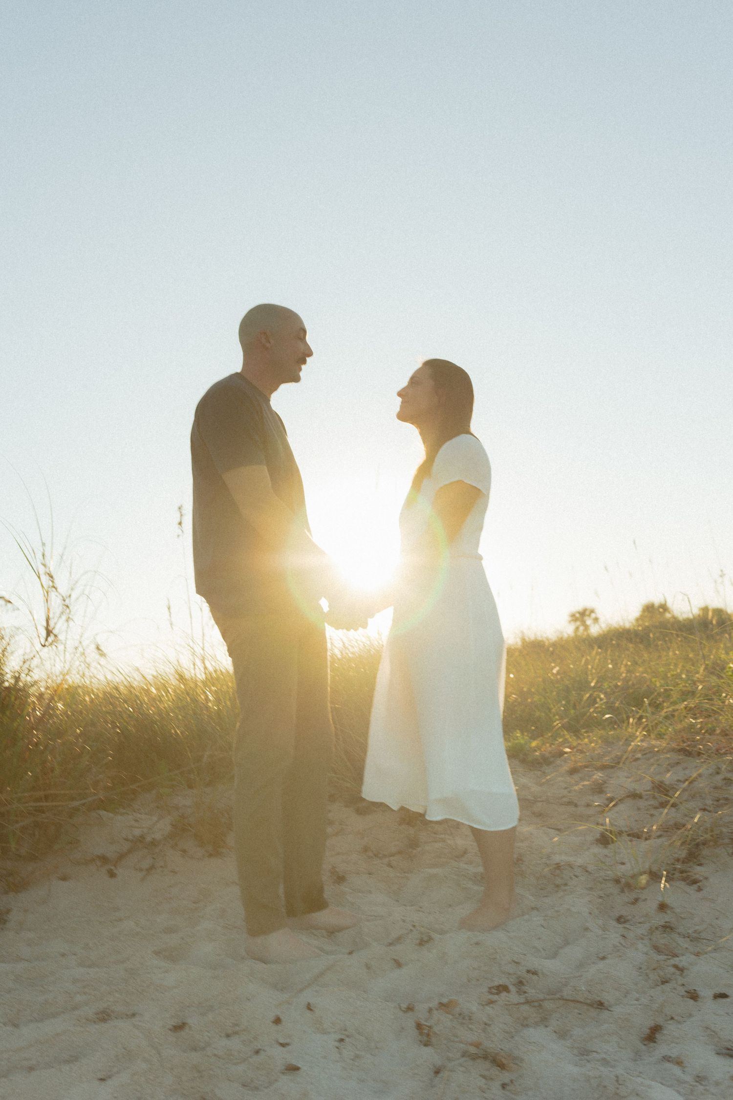 Couple holding hands facing each other on sandy beach at sunset with golden lens flare behind them.