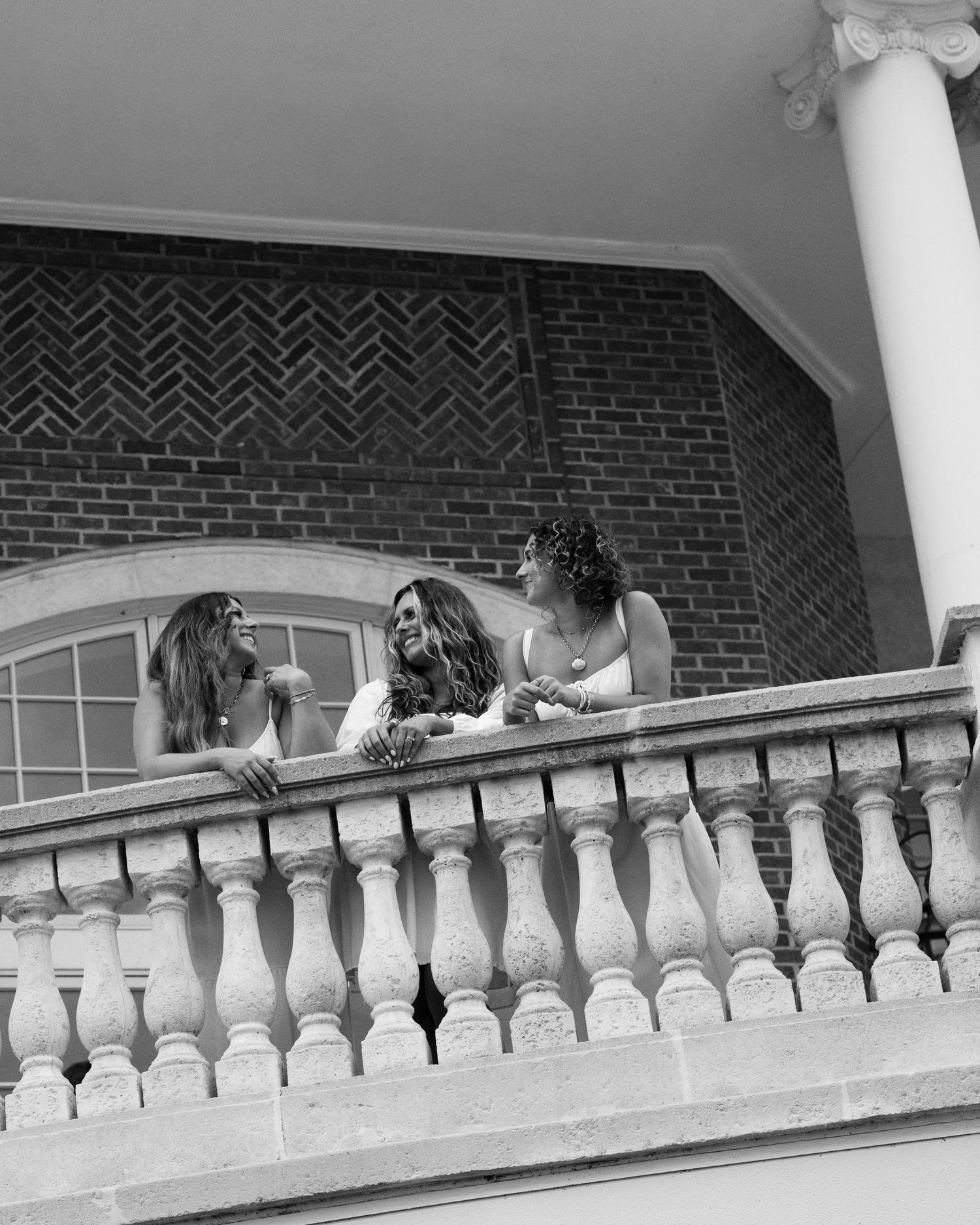 Black and white photo of three women laughing on a decorative stone balcony railing.