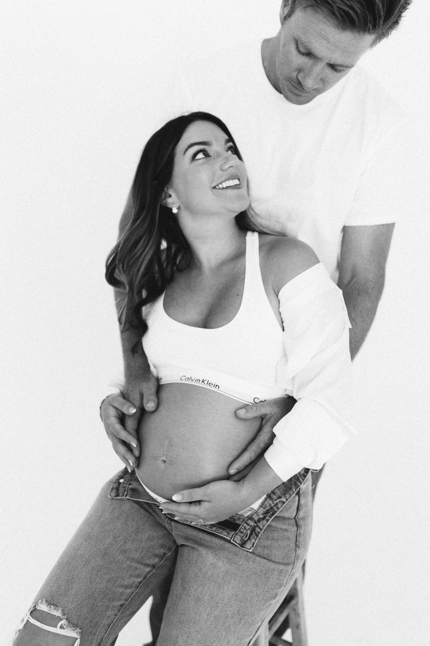 Black and white maternity photo of a smiling pregnant woman in Calvin Klein bralette with her partner.