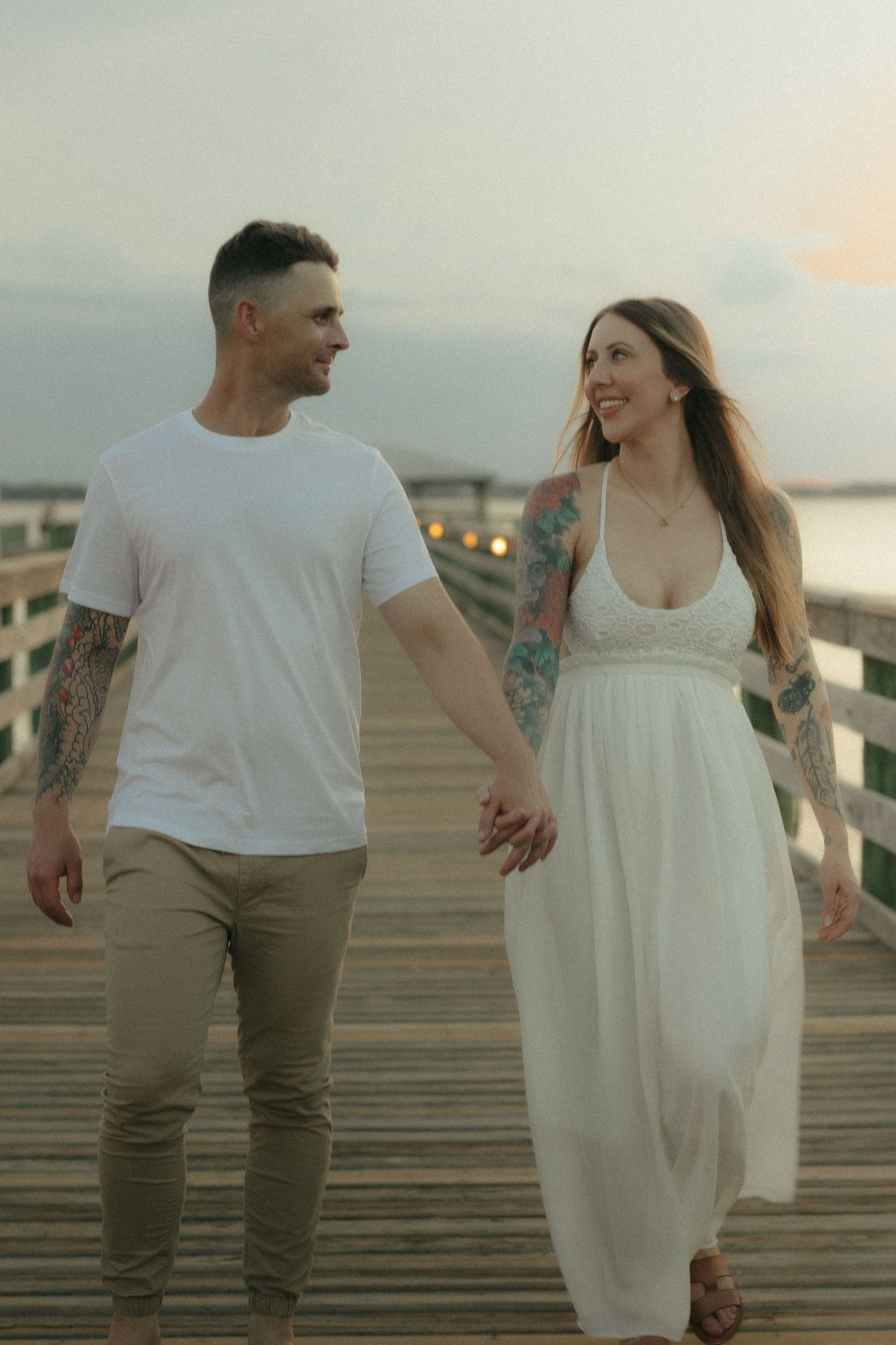 Smiling tattooed couple holding hands and walking together on a waterfront boardwalk at dusk.