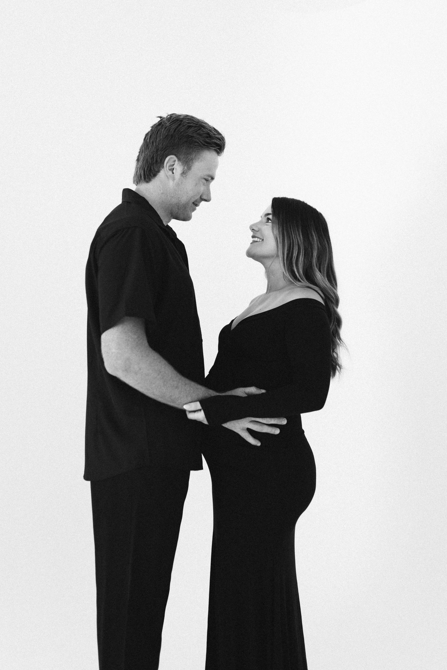 Black and white maternity photo of a smiling couple in black outfits embracing and gazing at each other.