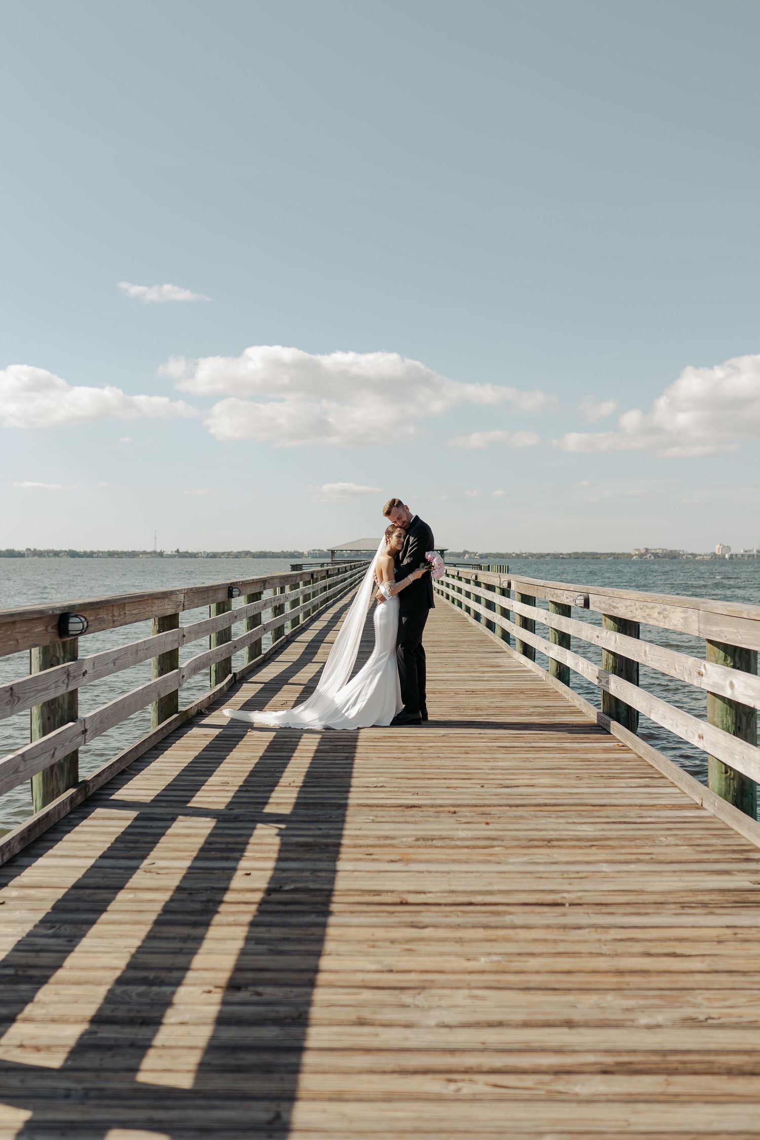 Bride and groom embrace on a long wooden pier overlooking calm blue water under a sunny sky.