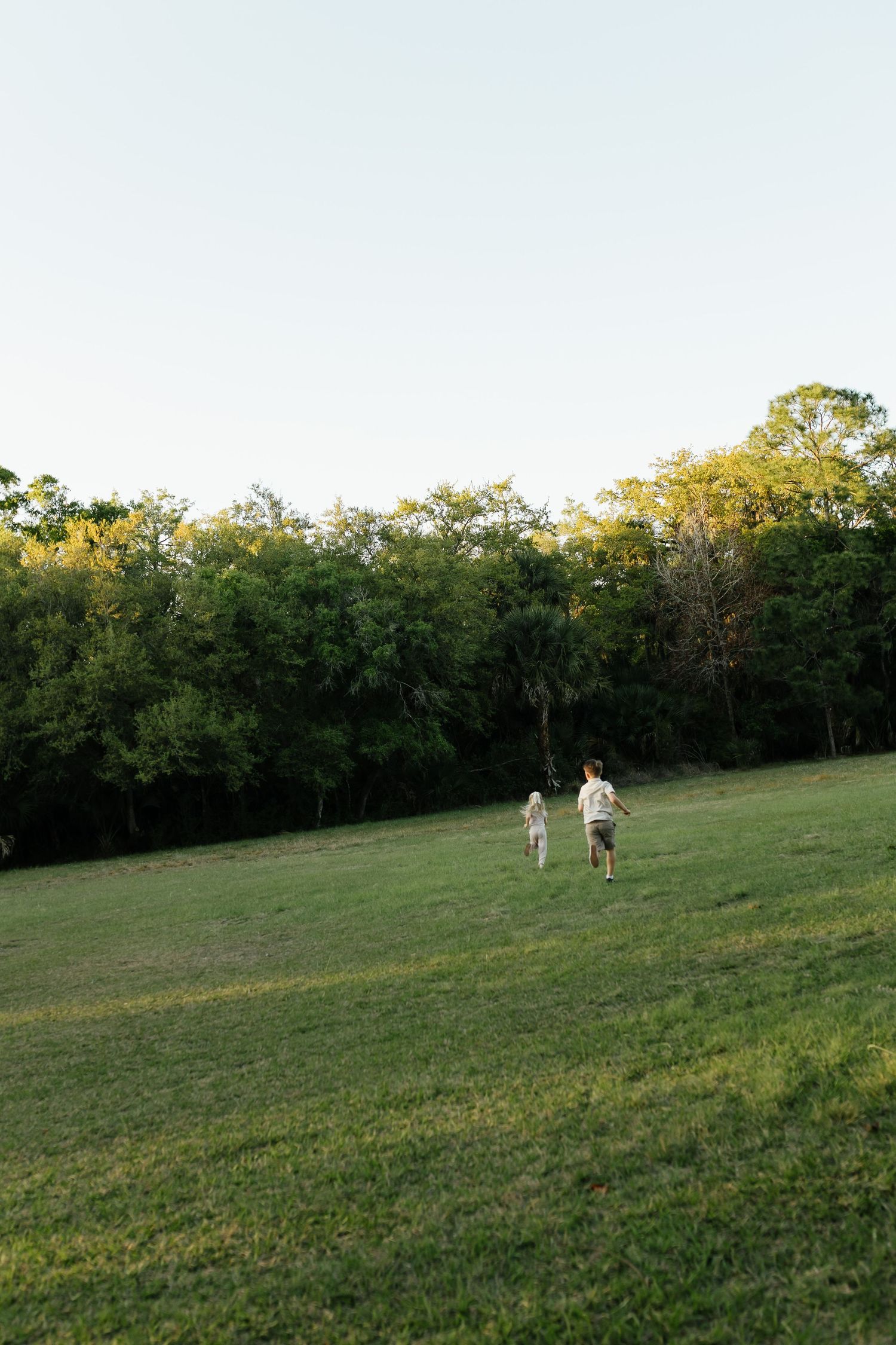 Two children running across a lush green field toward a dense tree line on a clear day.