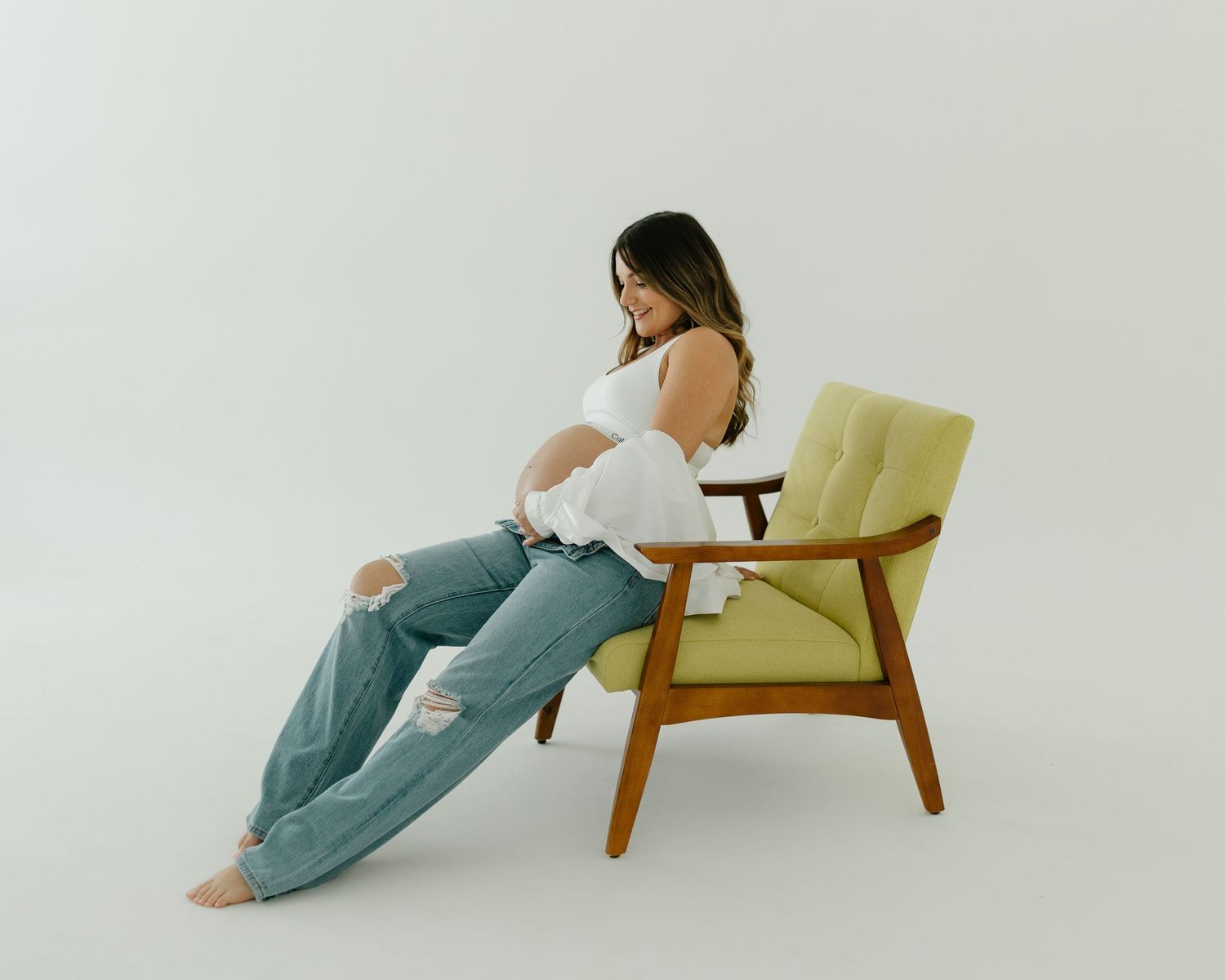 Pregnant woman in white top and ripped jeans sits on a yellow mid-century modern chair, cradling her baby bump.