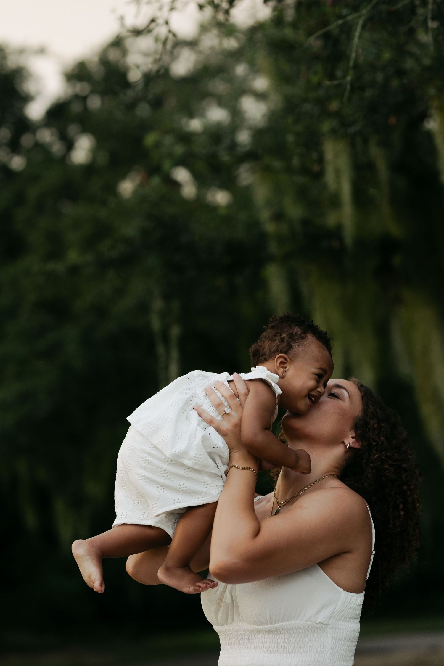 A loving mother kisses her baby while lifting her up outdoors surrounded by green trees.