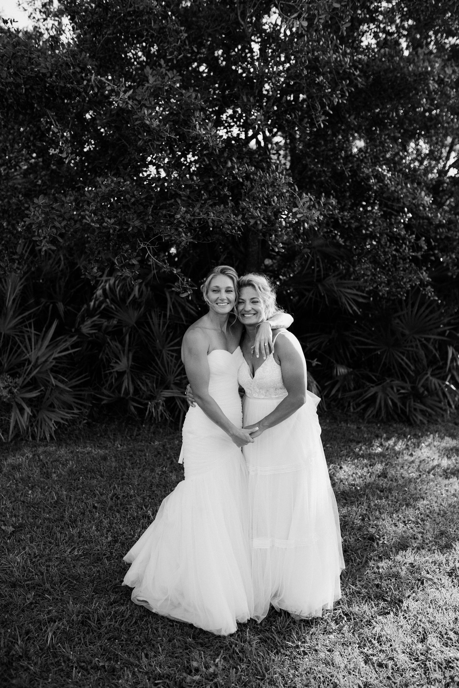 Two brides in white gowns pose together smiling in a black and white outdoor wedding portrait.