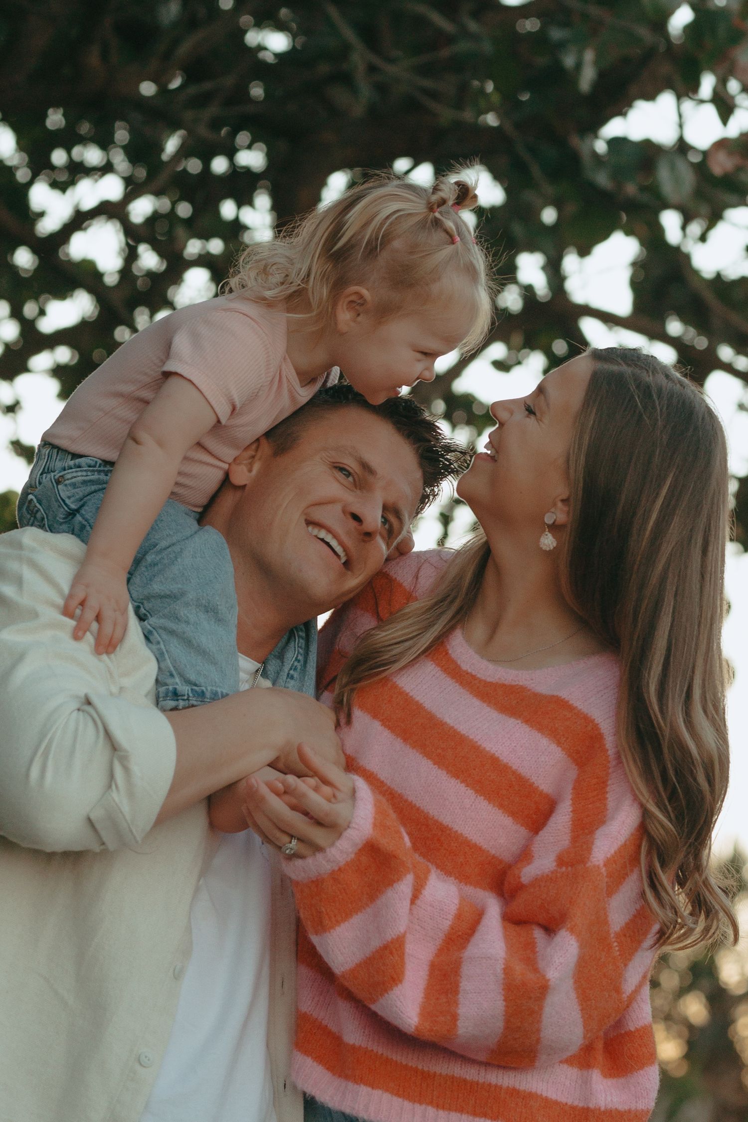 Happy family with laughing dad carrying toddler on shoulders while mom smiles outdoors under tree.