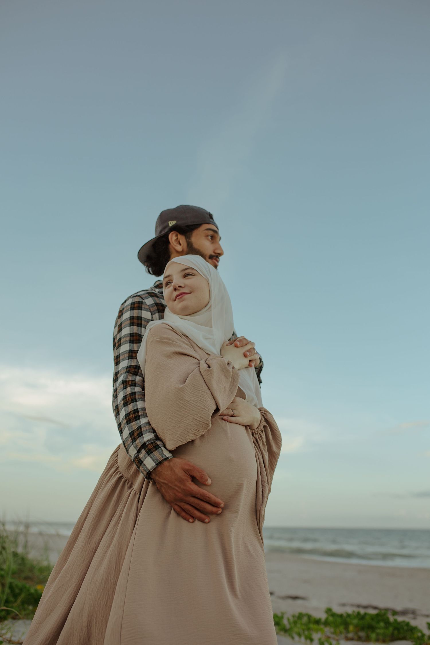 A Muslim couple embracing on a beach at dusk, the woman wearing a hijab and visibly pregnant, both looking peacefully into the distance.
