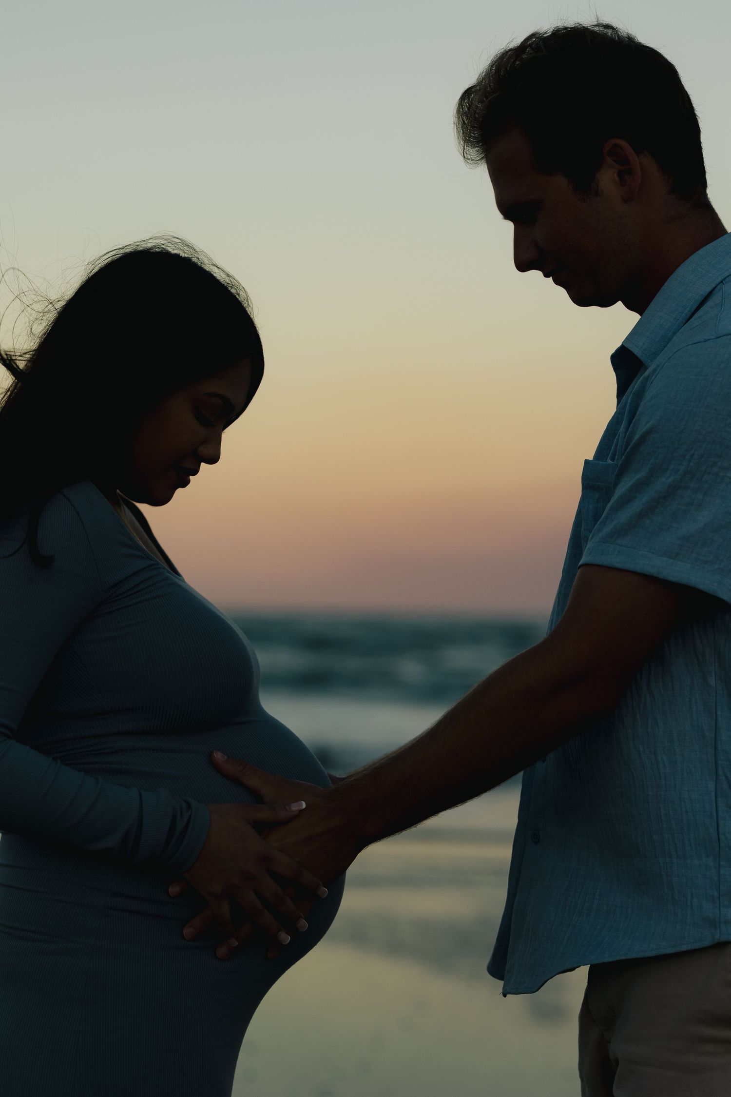 Silhouette of a pregnant couple at sunset on the beach, both tenderly touching the baby bump.