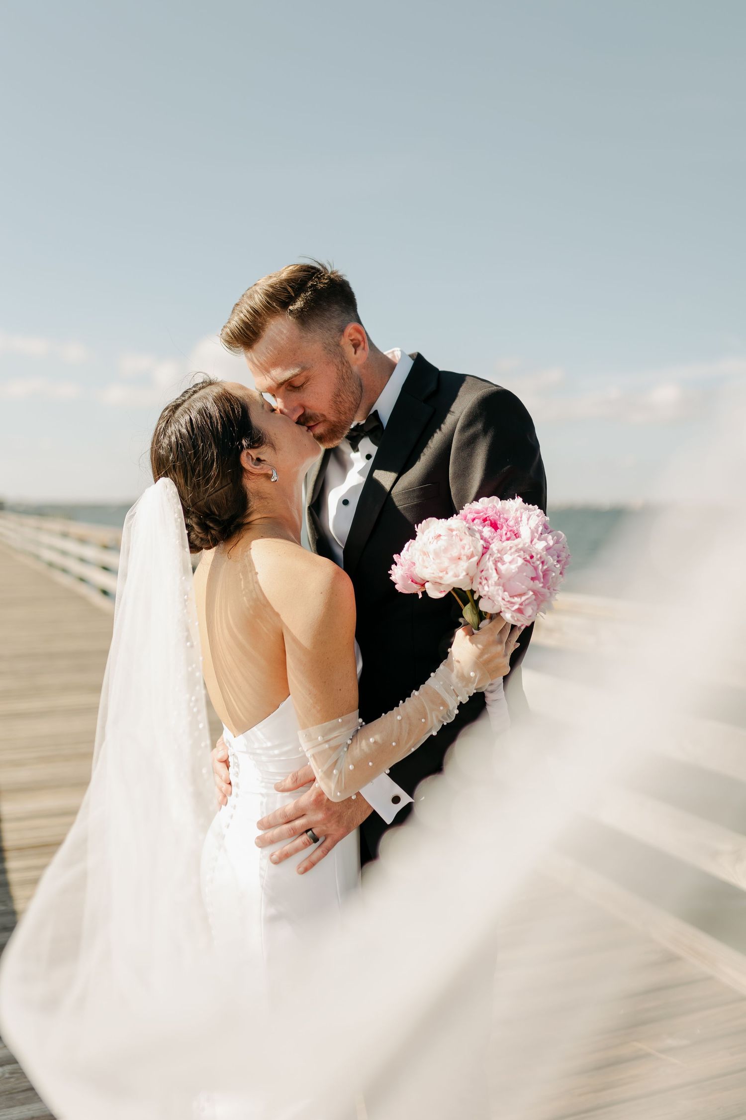 Bride and groom sharing a romantic kiss on a waterfront pier with veil blowing in the wind.
