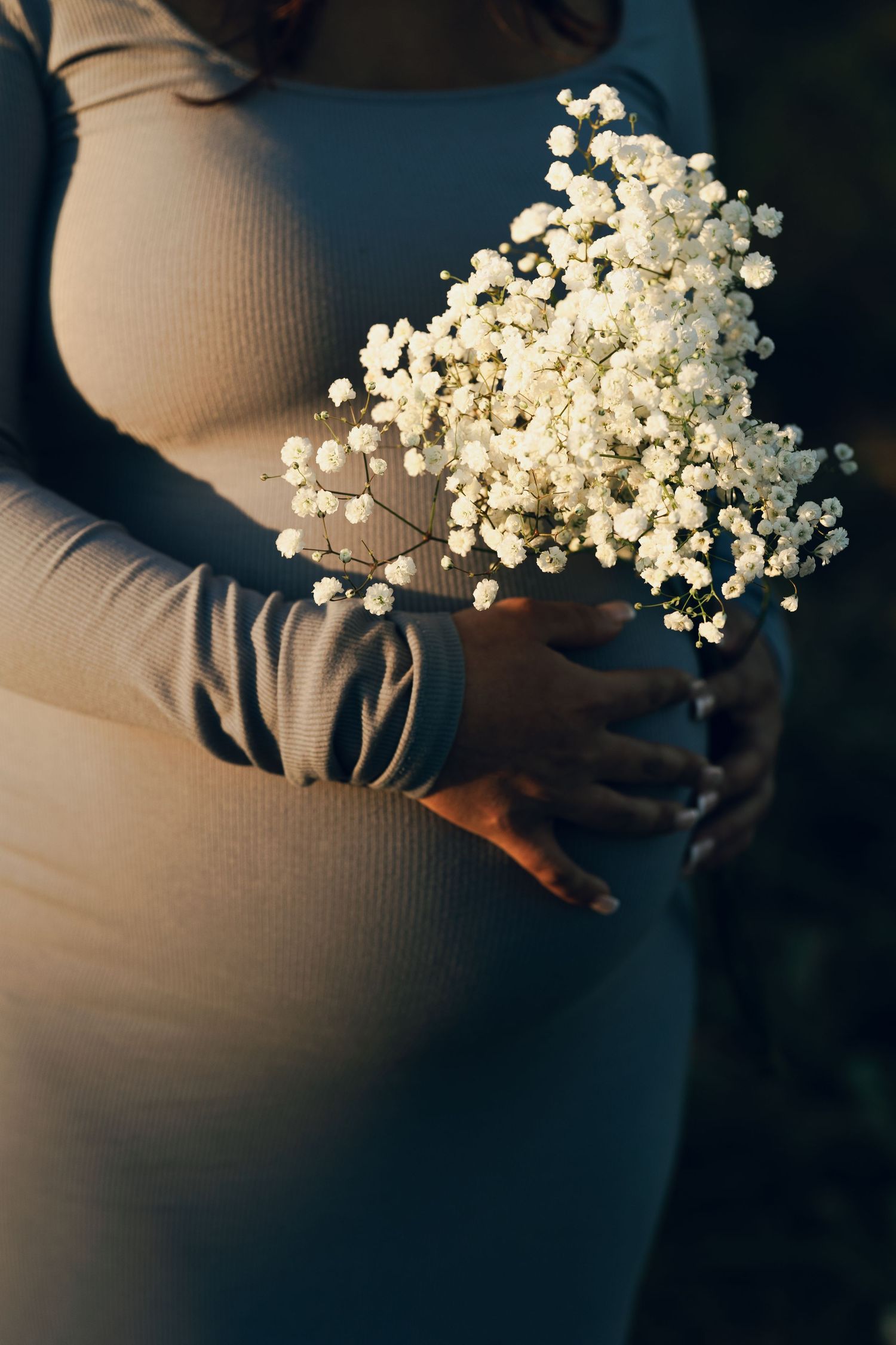 Close-up of a pregnant woman in a beige dress holding white baby's breath flowers over her bump.