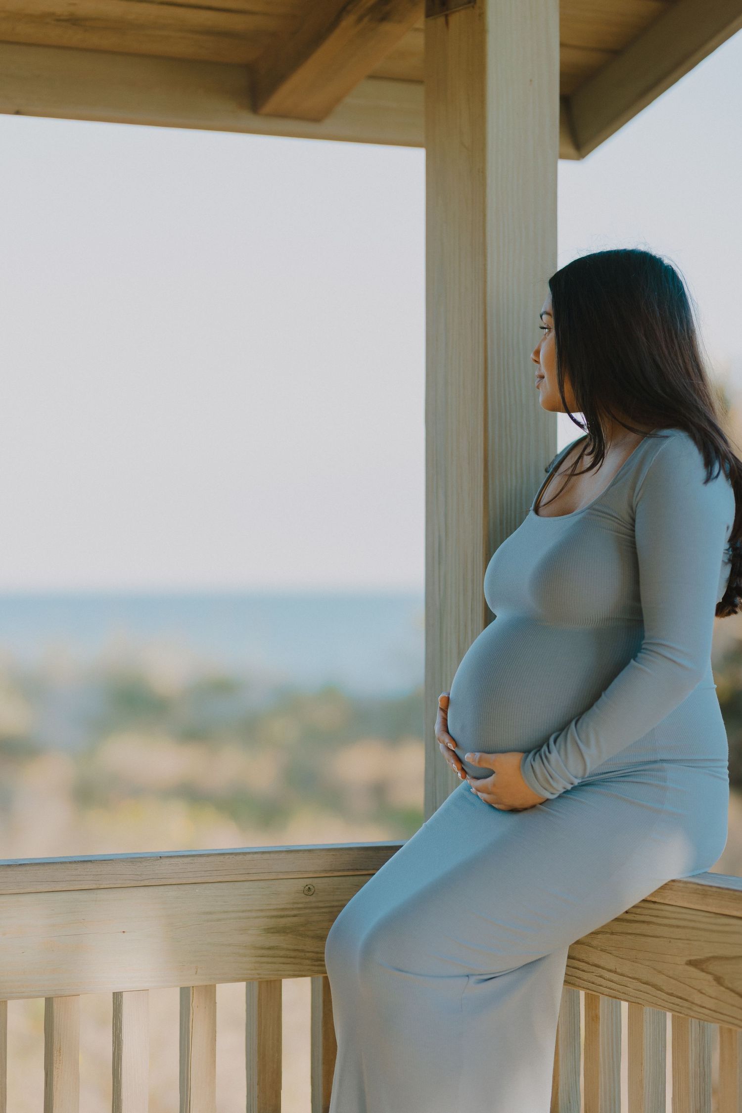 Pregnant woman in a blue dress sitting on a porch railing overlooking a scenic outdoor view.