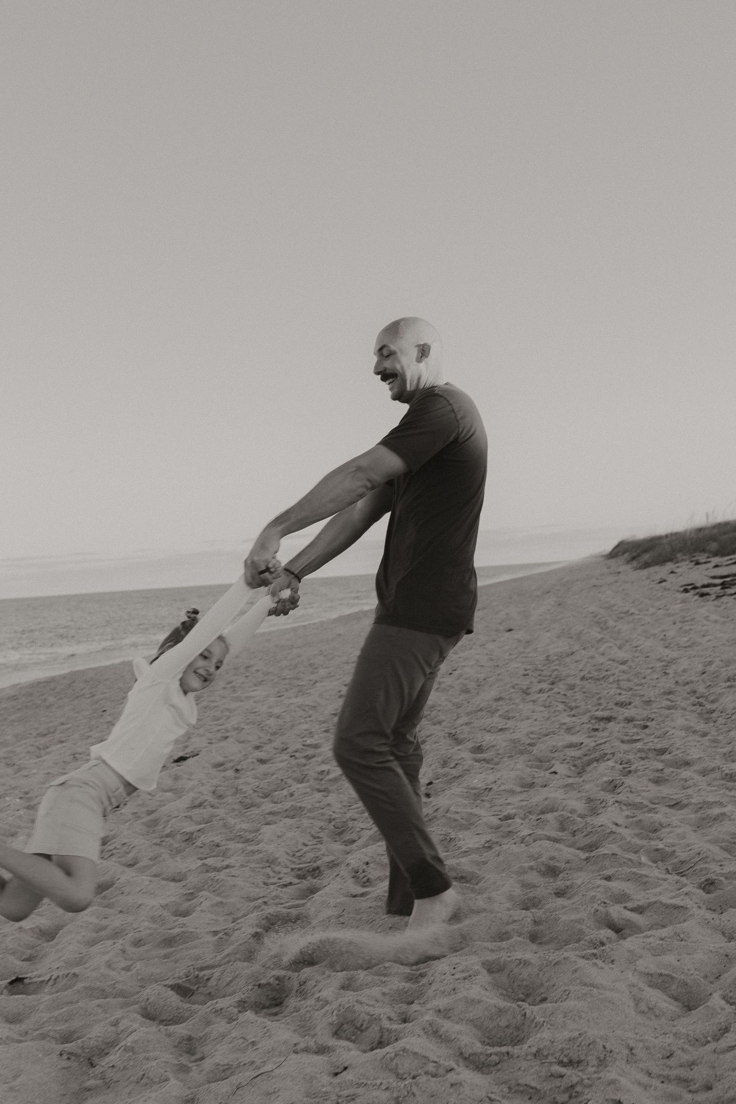 Black and white photo of bald father joyfully spinning young child by hands on sandy beach.