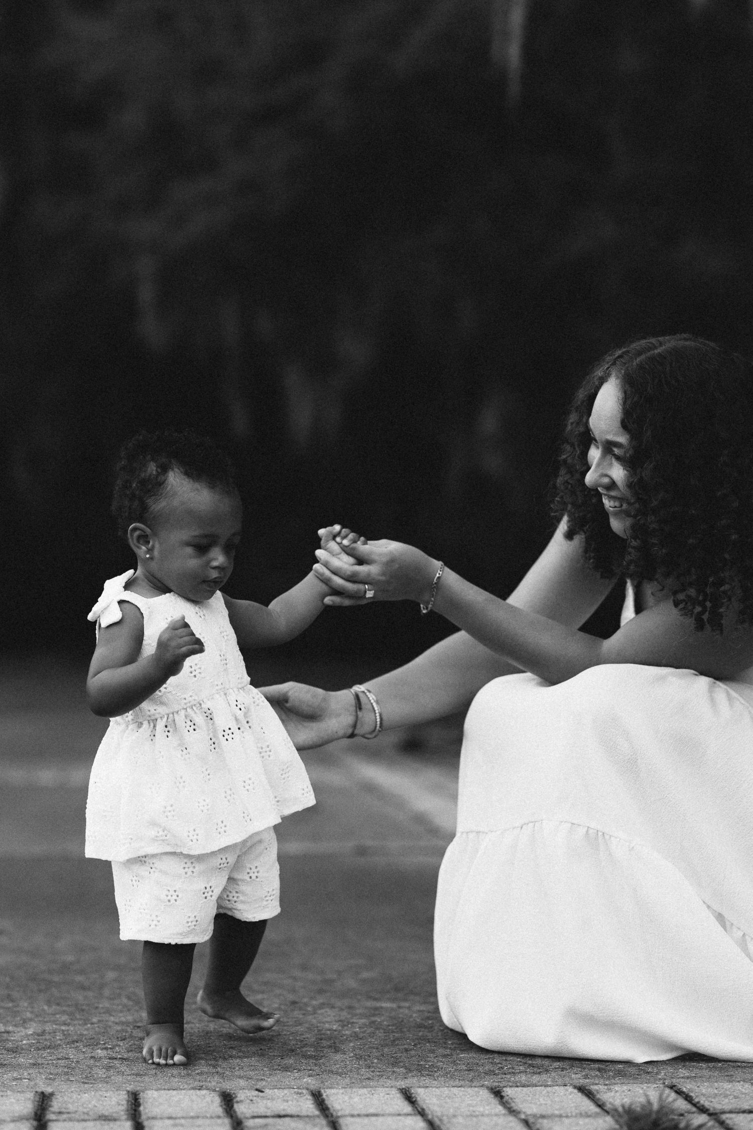 Black and white image of a mother and toddler girl holding hands and interacting on a brick path.
