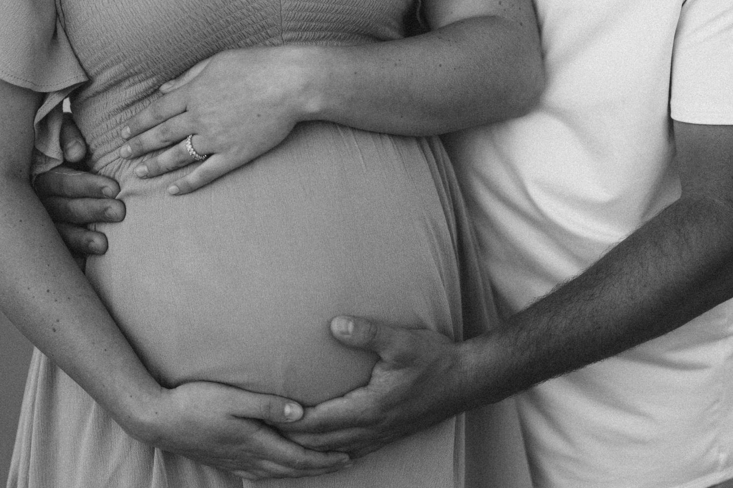A black and white close-up of a pregnant couple tenderly cradling a baby bump together.