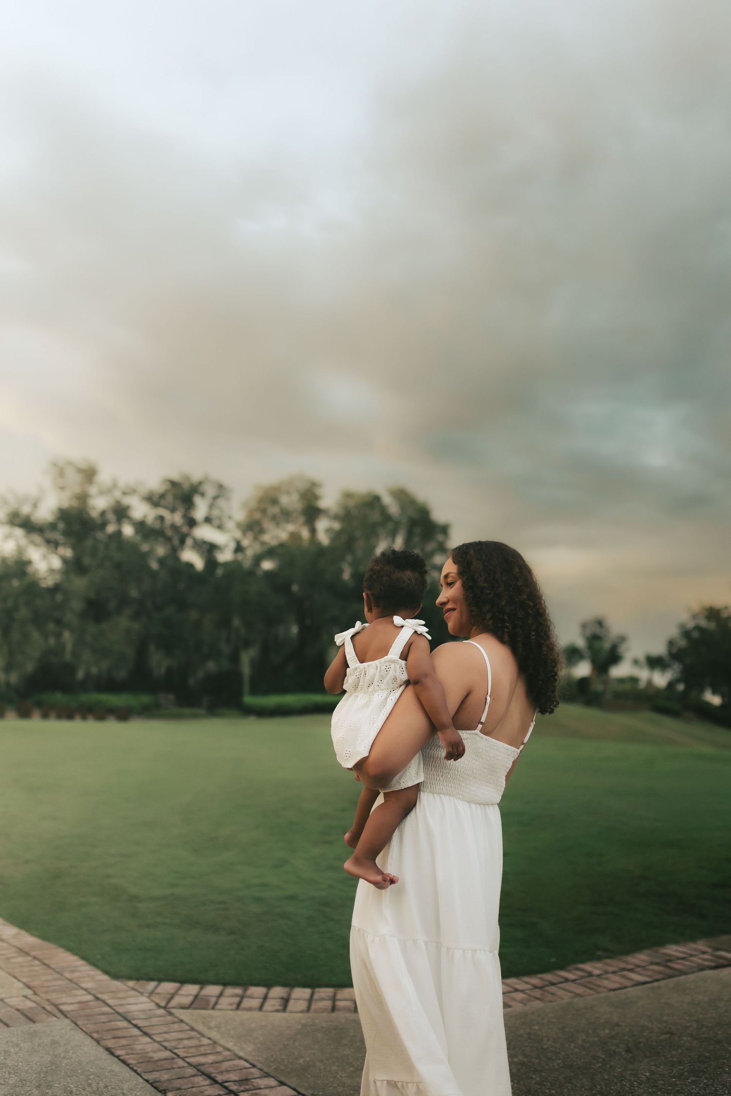 A mother in a white dress holds her baby girl outdoors on a cloudy evening near a lush green lawn.