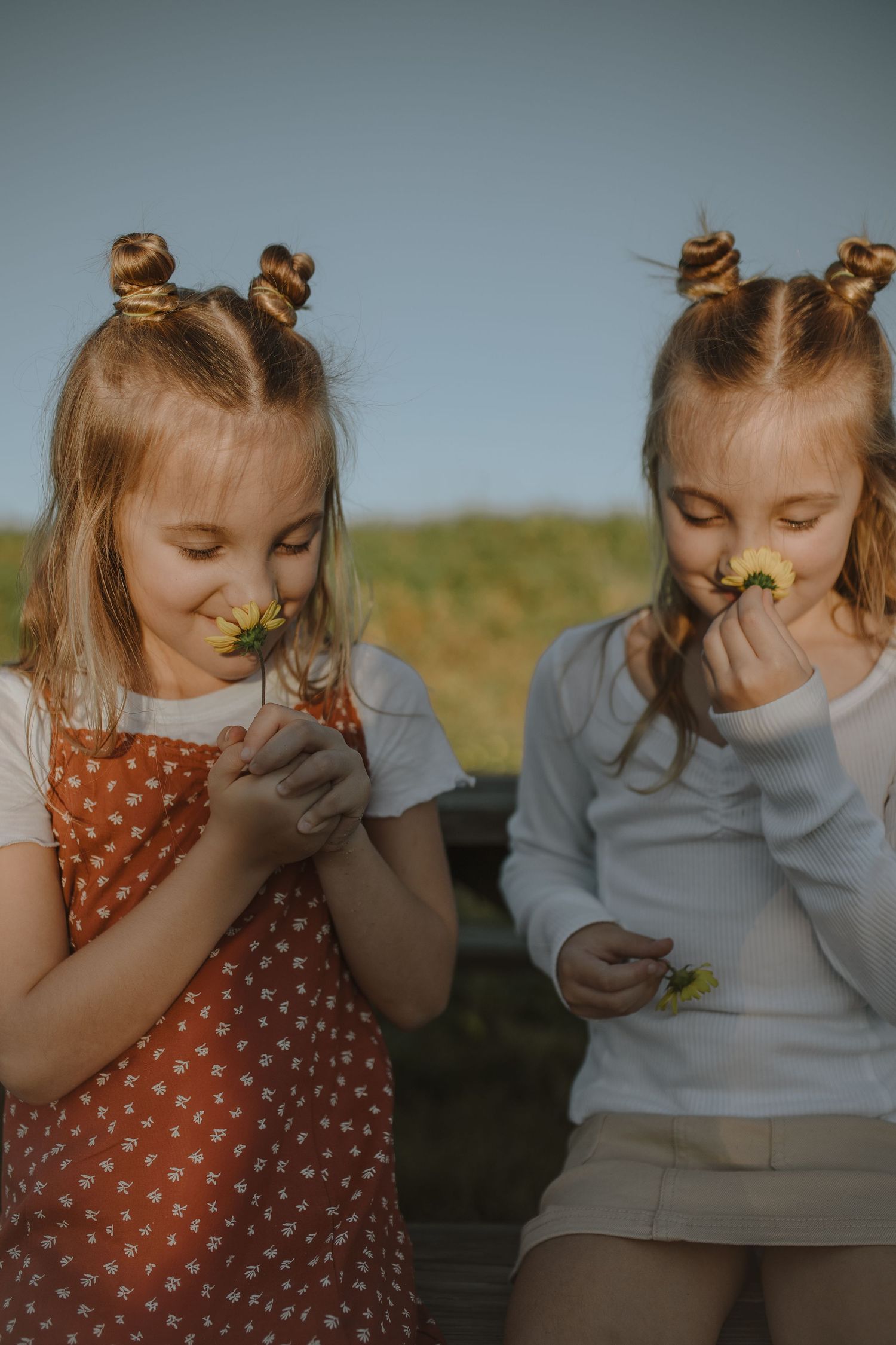 Two blonde girls with bun hairstyles smelling yellow wildflowers outdoors in warm golden sunlight.