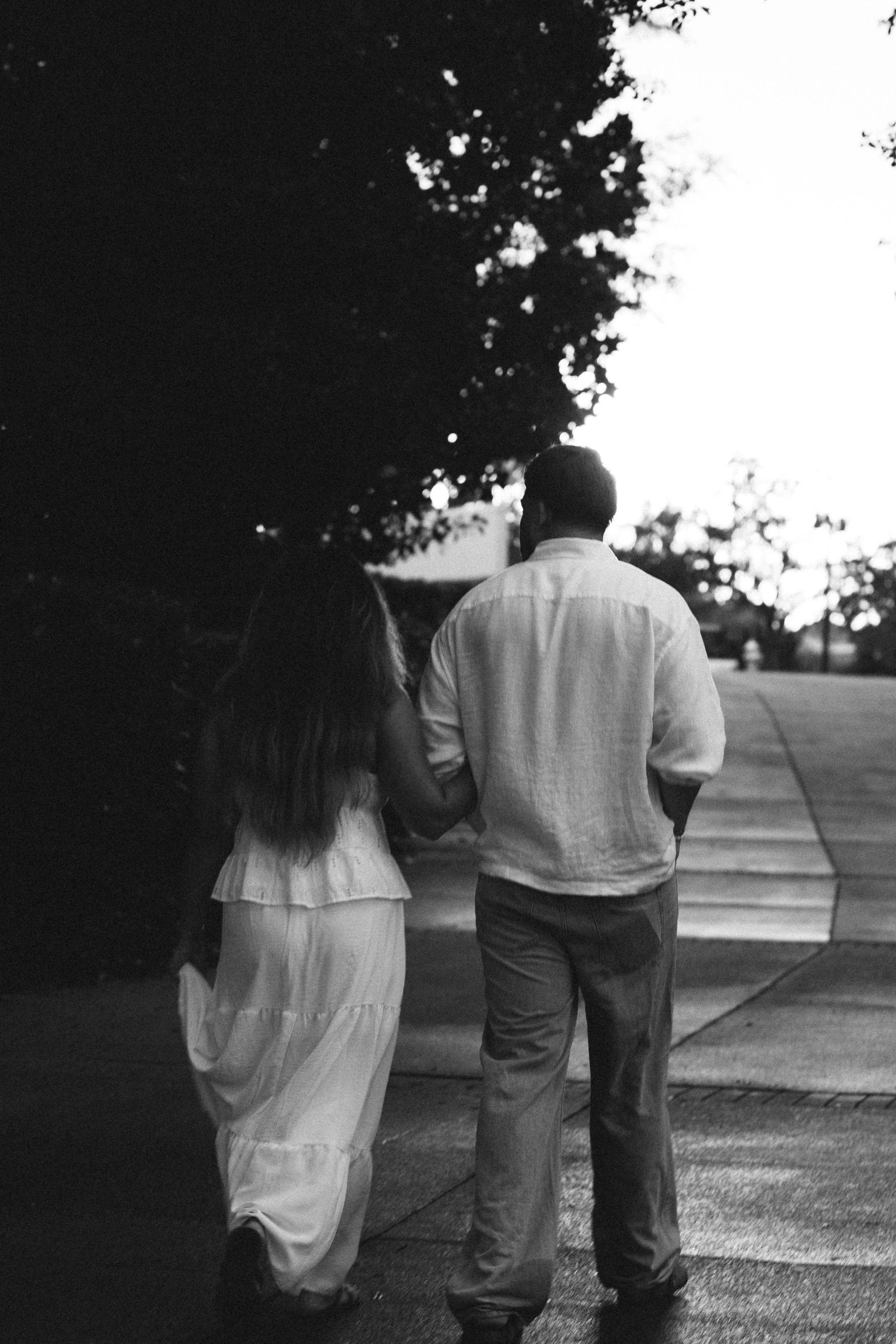 Black and white photo of a couple in white holding hands while walking along a tree-lined path.