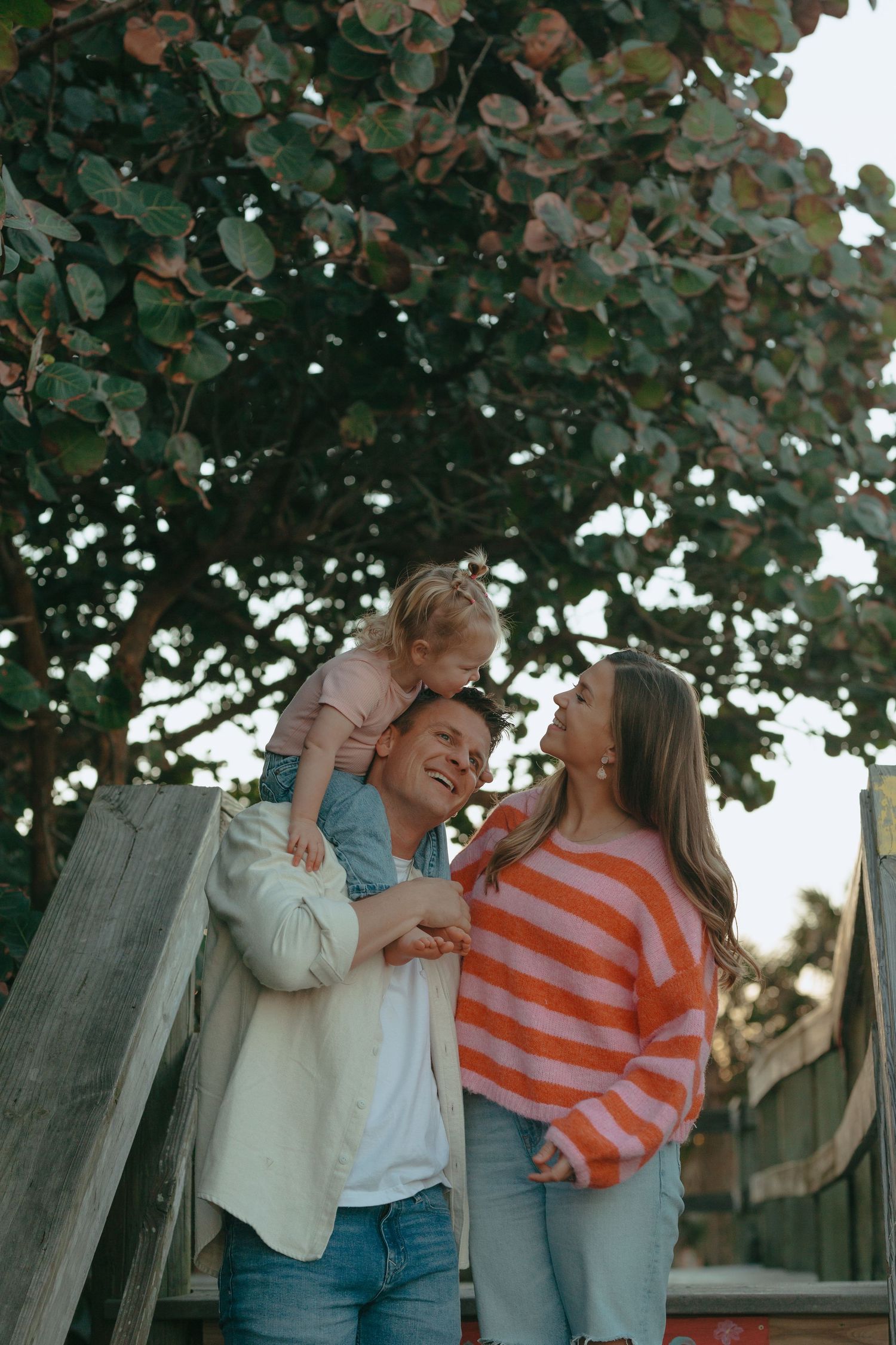 Joyful family with toddler on dad's shoulders under a lush green tree.