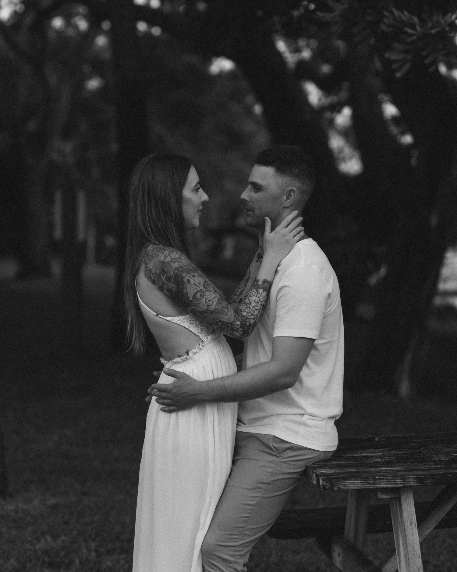 Black and white intimate portrait of a tattooed couple embracing tenderly in an outdoor park setting.