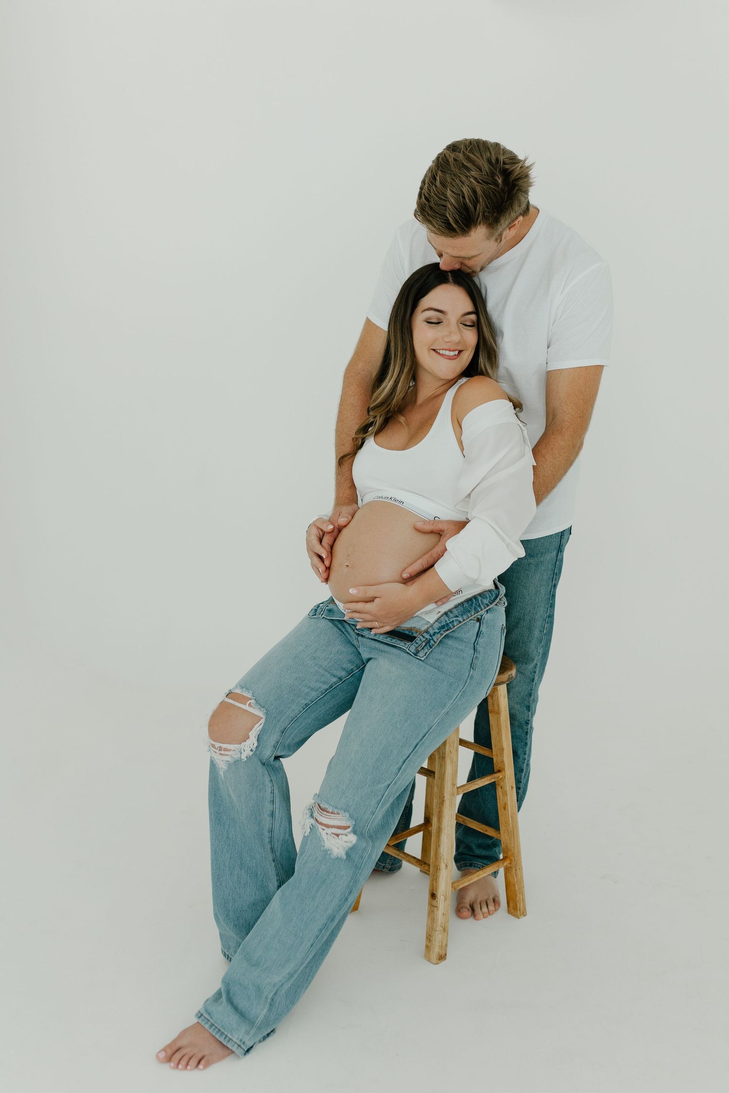 Couple in white tops and jeans posing for a maternity photo, man kissing womans head as she sits.