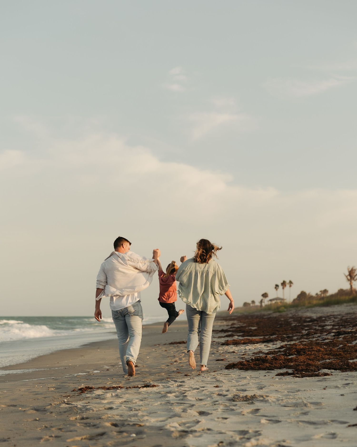 Family walking on beach at sunset, parents swinging child between them with ocean waves and palm trees in background.