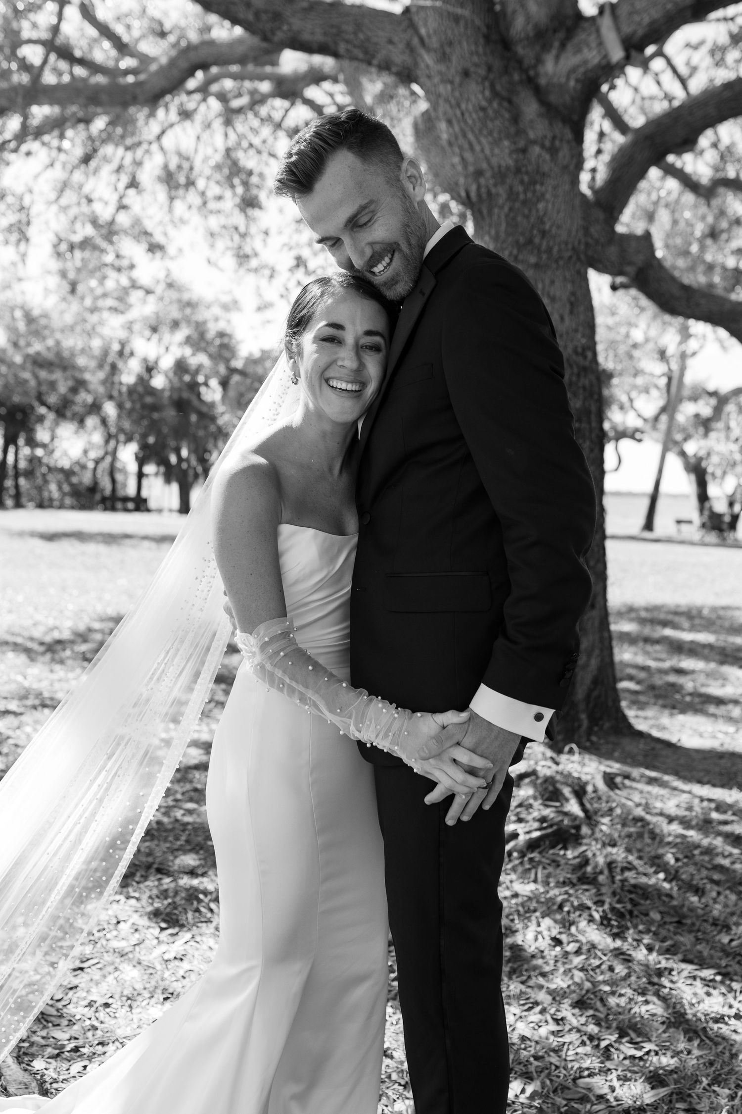 Black and white photo of smiling bride and groom embracing outdoors under a large tree on their wedding day.