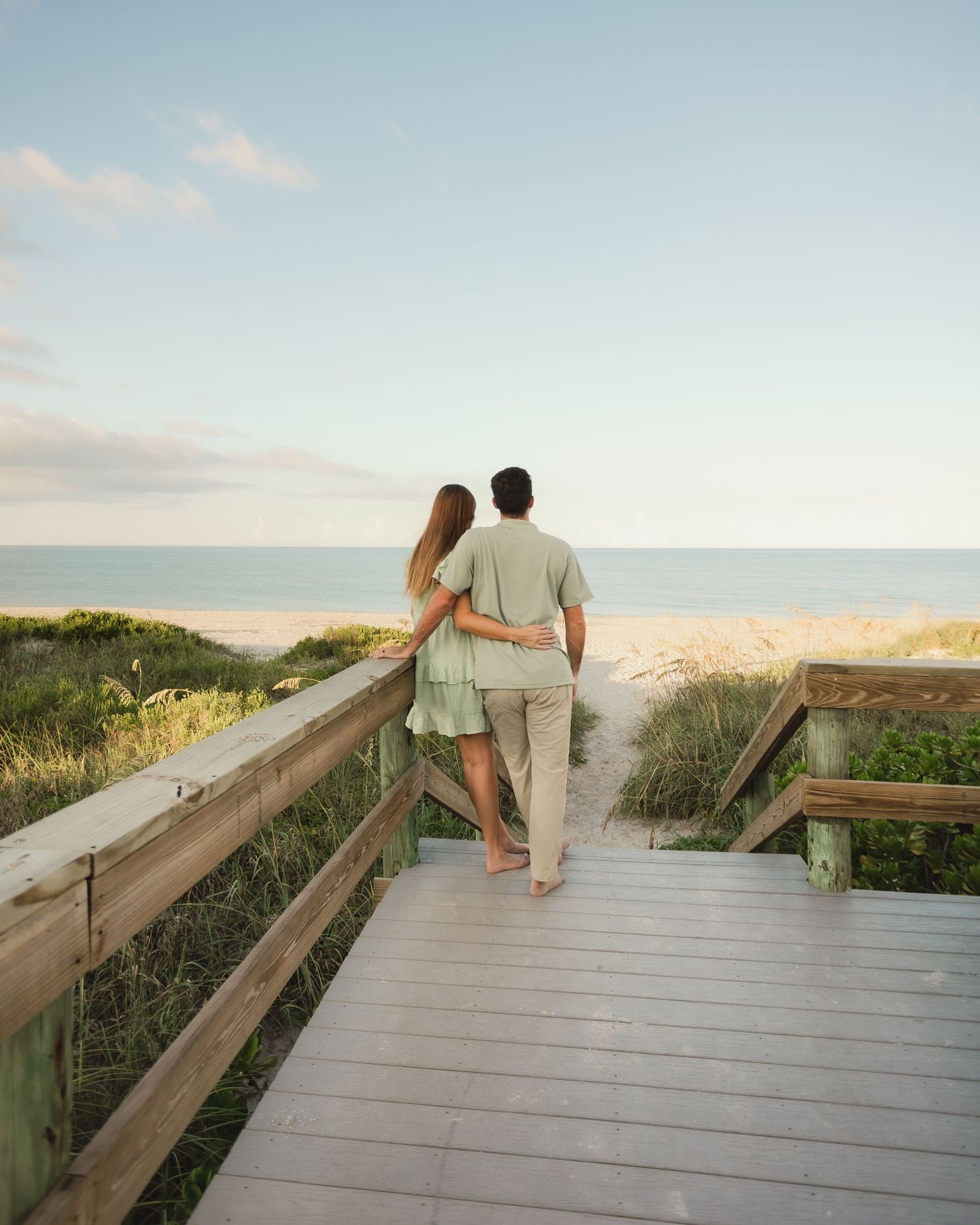 Couple standing on a wooden boardwalk overlooking a serene beach at sunset, embracing each other.