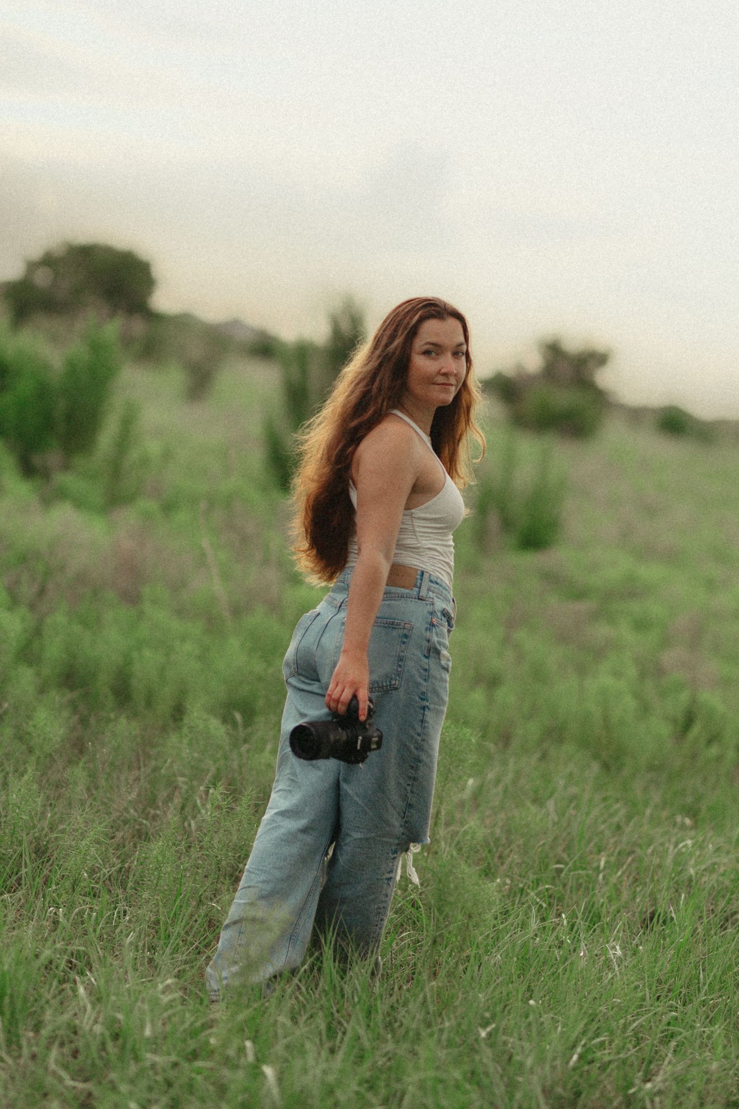 Smiling young woman with long red hair holding a camera in a green meadow, wearing white crop top and jeans.