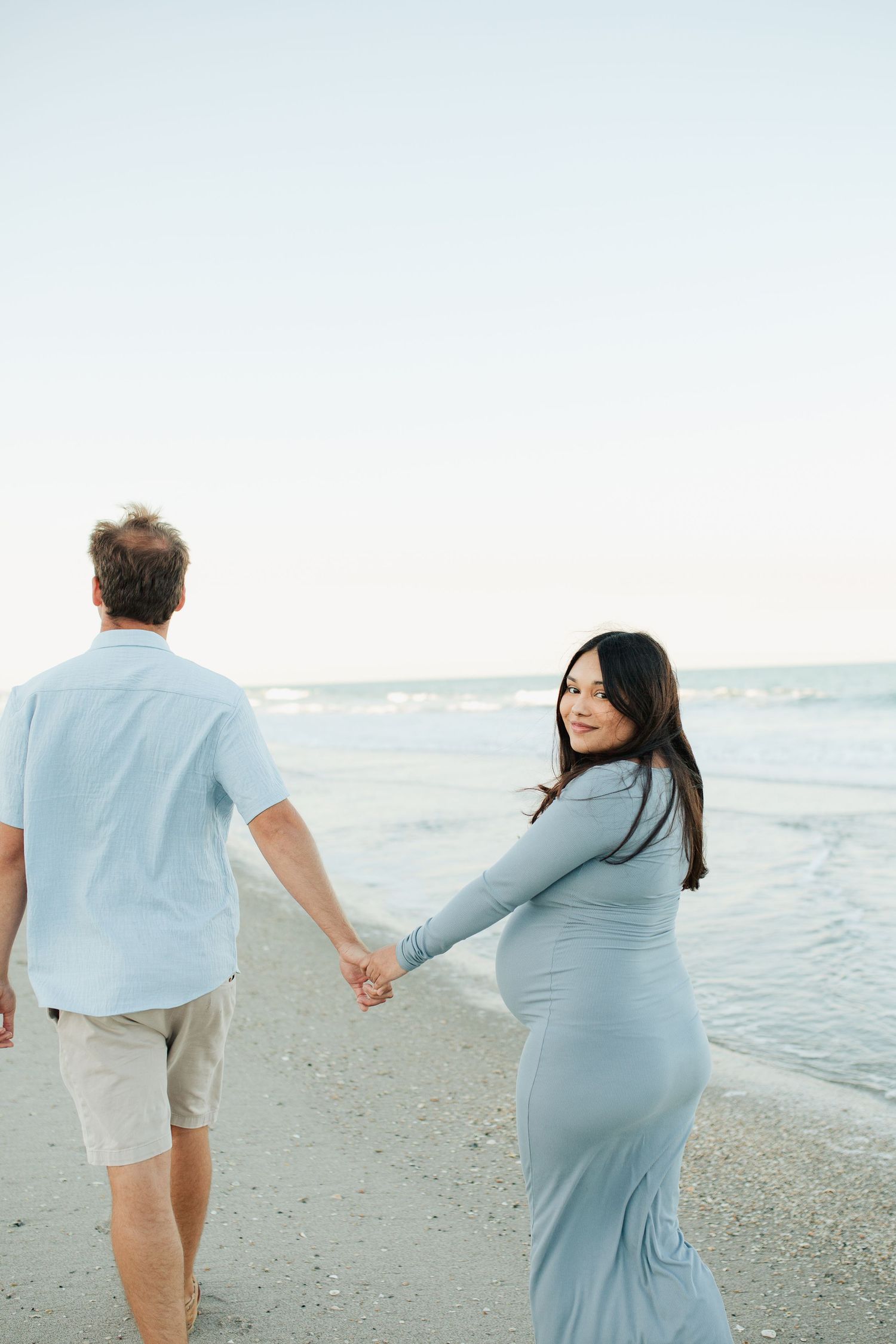 Pregnant woman in a blue dress holding hands with her partner while walking along a serene beach.