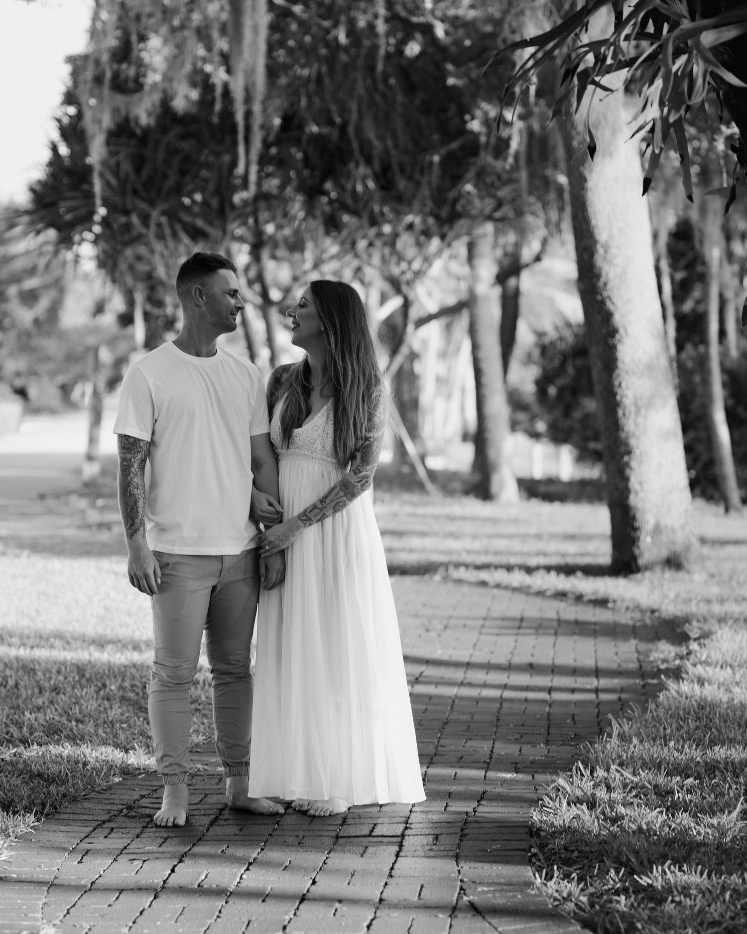 Black and white photo of tattooed couple holding hands on a tree-lined brick pathway.