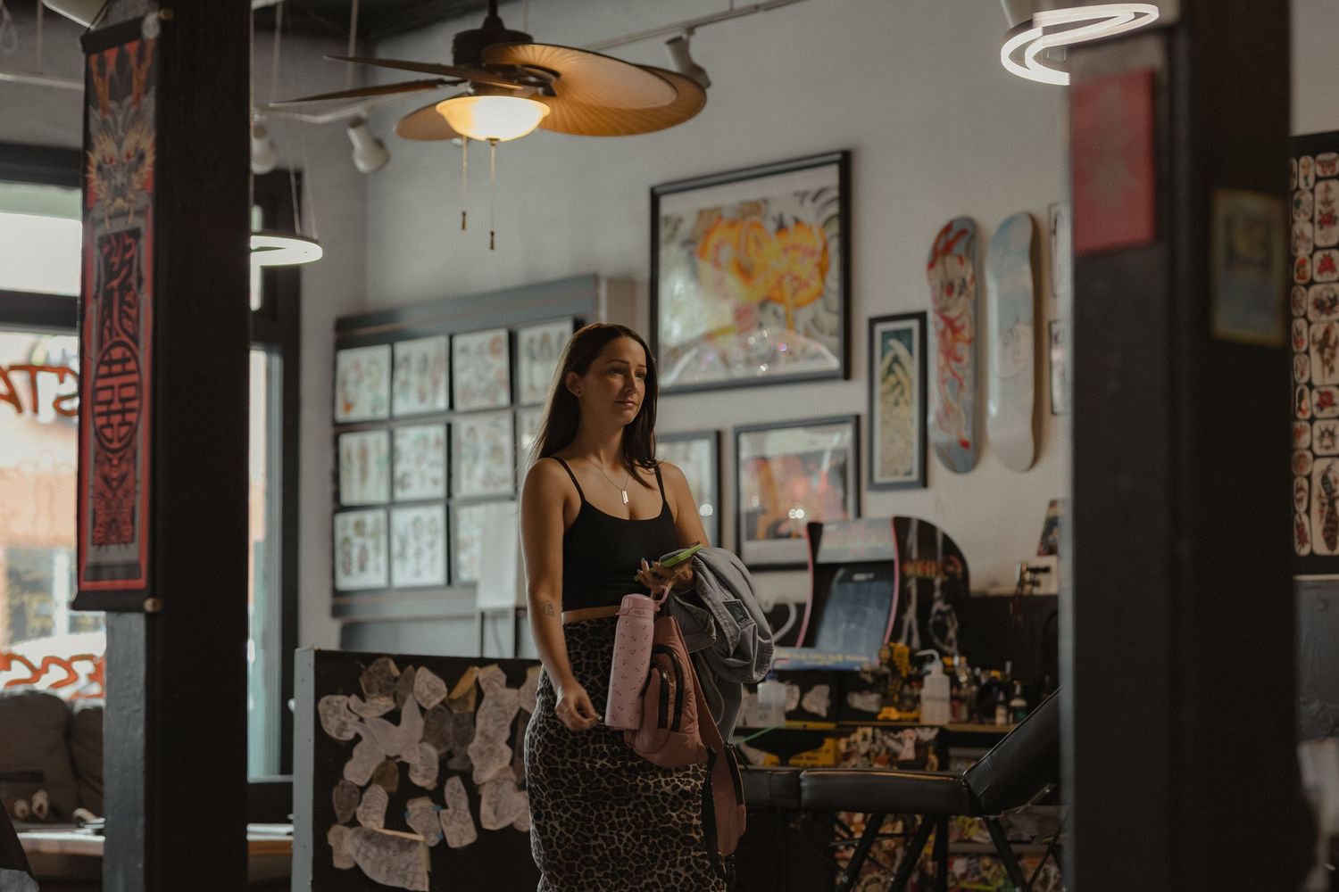 Young woman with tattoos standing inside a stylish tattoo parlor holding a backpack.