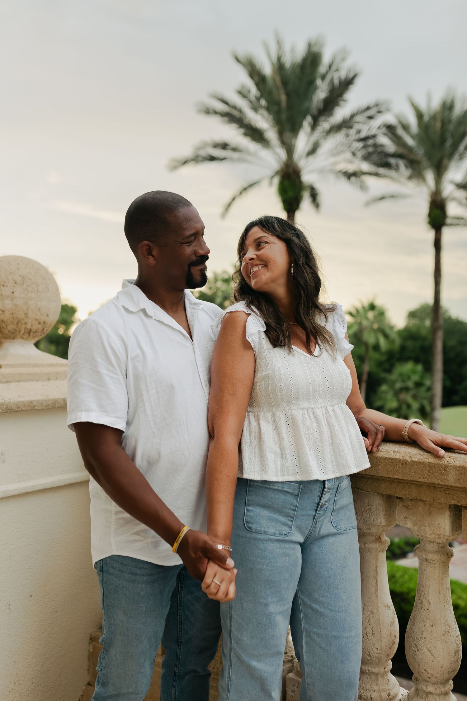 Couple in white tops and jeans holding hands on a stone balcony with palm trees.